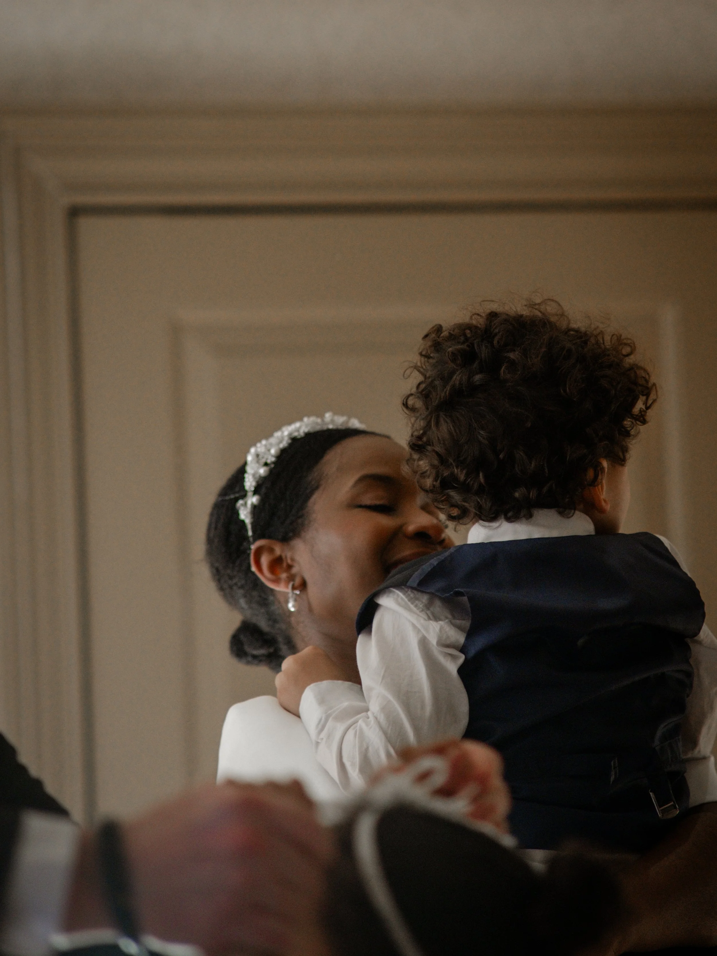 A woman with a pearl headband is hugging and smiling at a young boy with curly hair, both dressed formally, in an indoor setting.