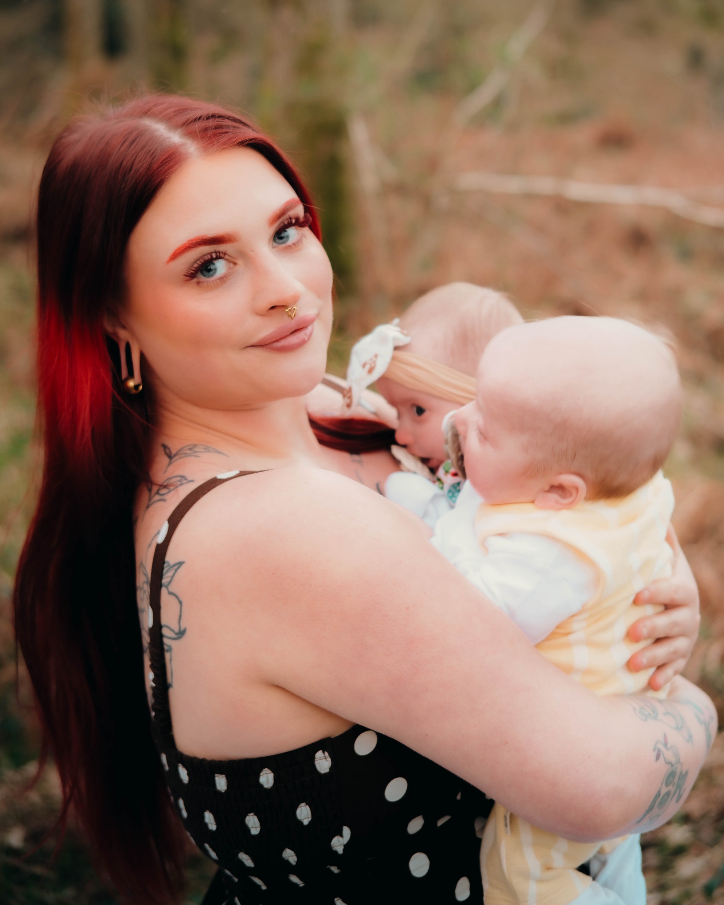 A woman with long red hair, blue eyes, and tattoos on her arm holds two babies, one with a white bow in its hair and the other with light blonde hair, outdoors with blurred trees in the background.