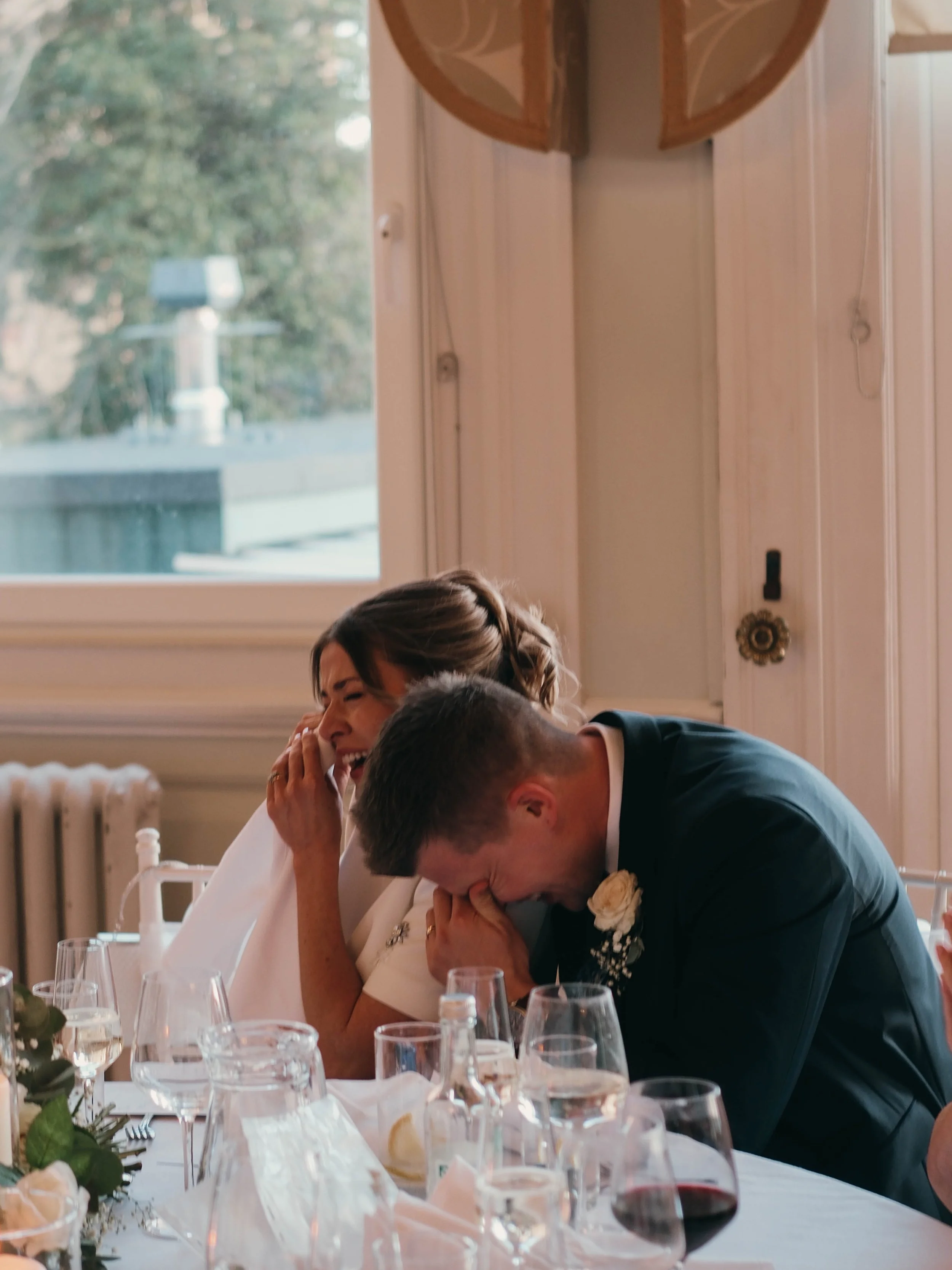 A woman and a man are both crying and leaning into each other at a celebration dinner table, with glasses and table decor visible in the foreground.