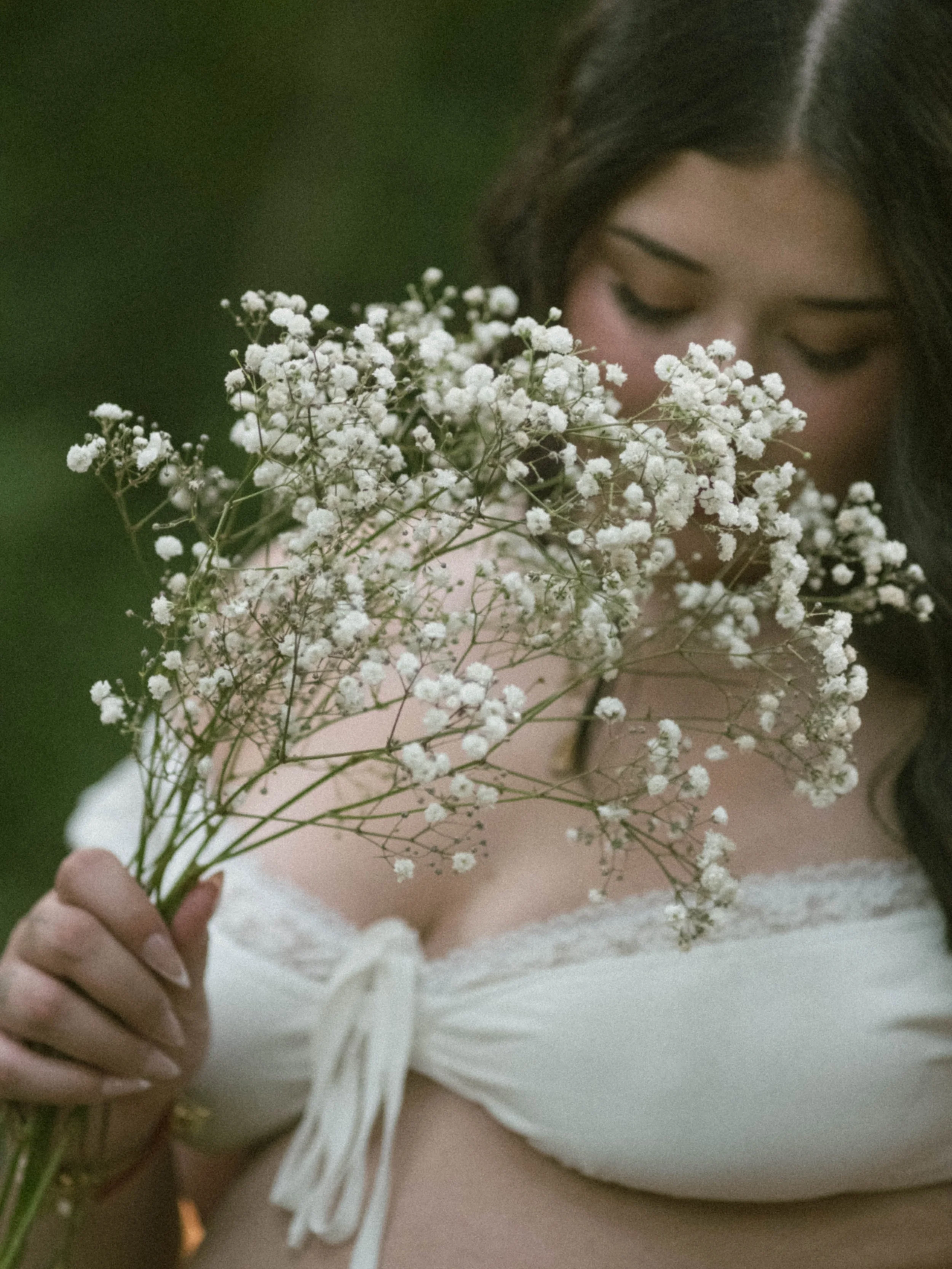 A person holding a small bouquet of white baby's breath flowers, with their face softly visible in the background and wearing a white lace dress.