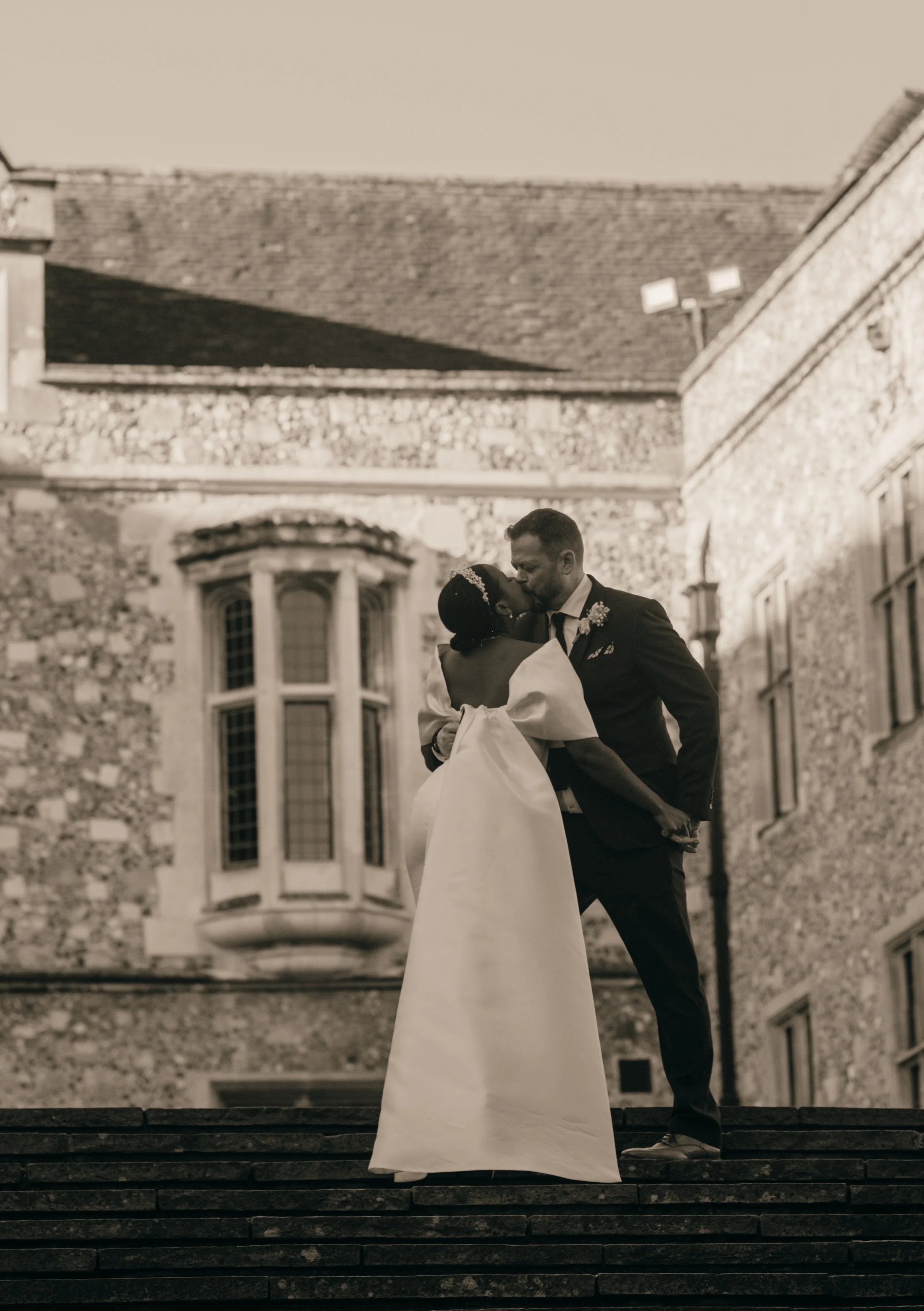 A newlywed couple sharing a kiss while standing on steps outside a historic stone building with large windows, during sunset.