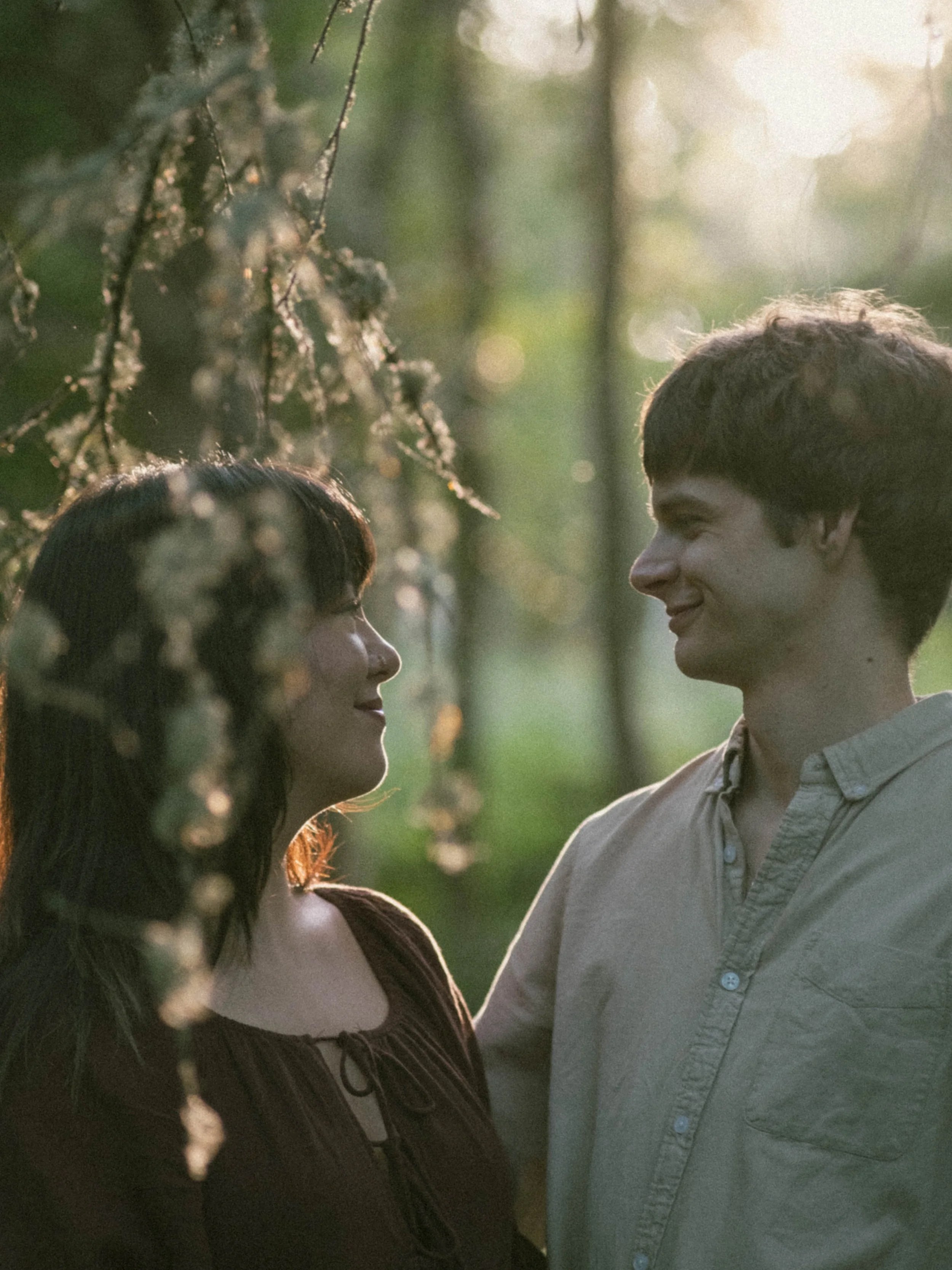 A young man and woman stand close to each other outdoors in a forest setting, gazing at each other, with sunlight filtering through the trees.