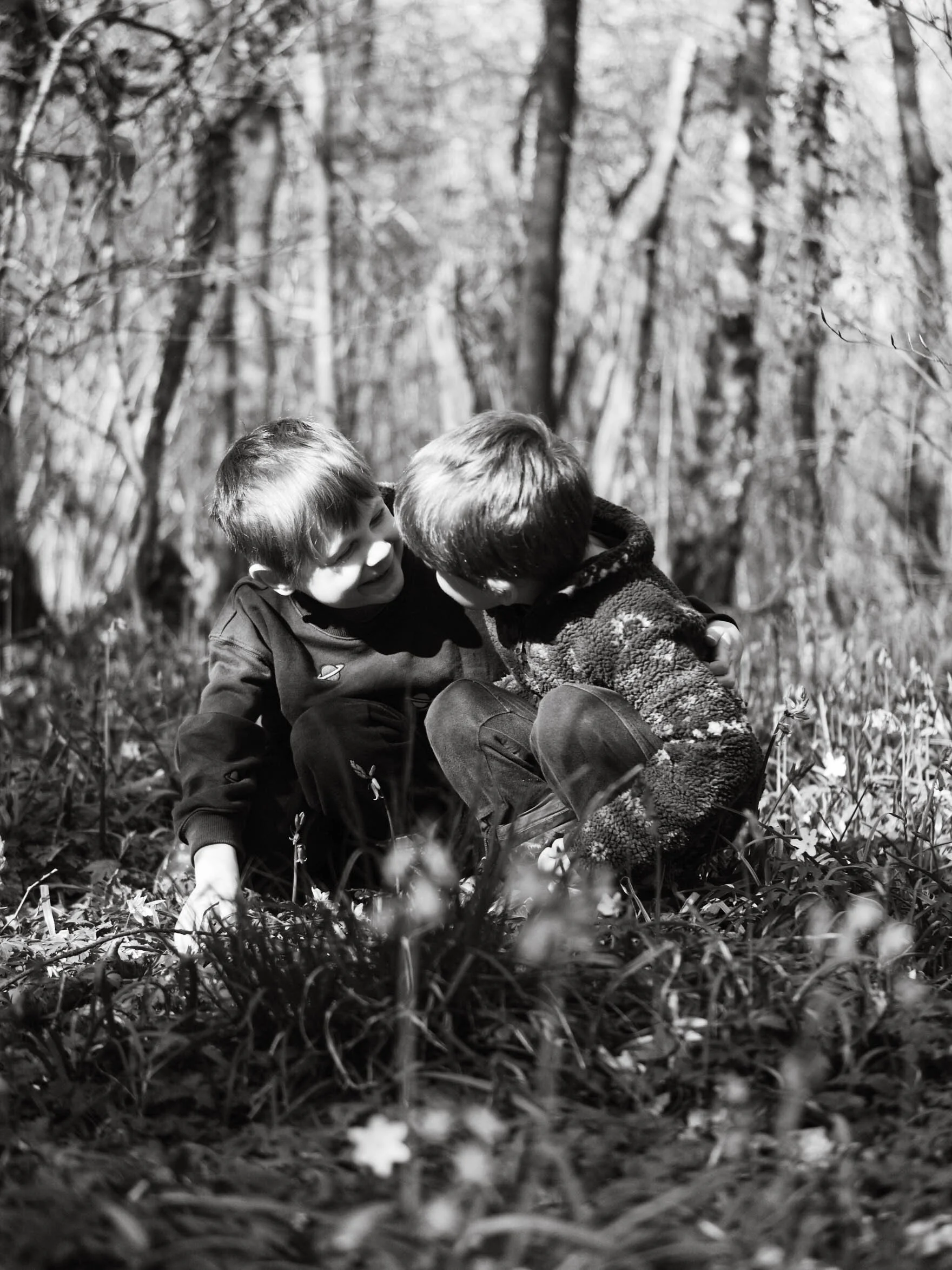 Two children playing together in a wooded area, smiling and leaning close to each other.