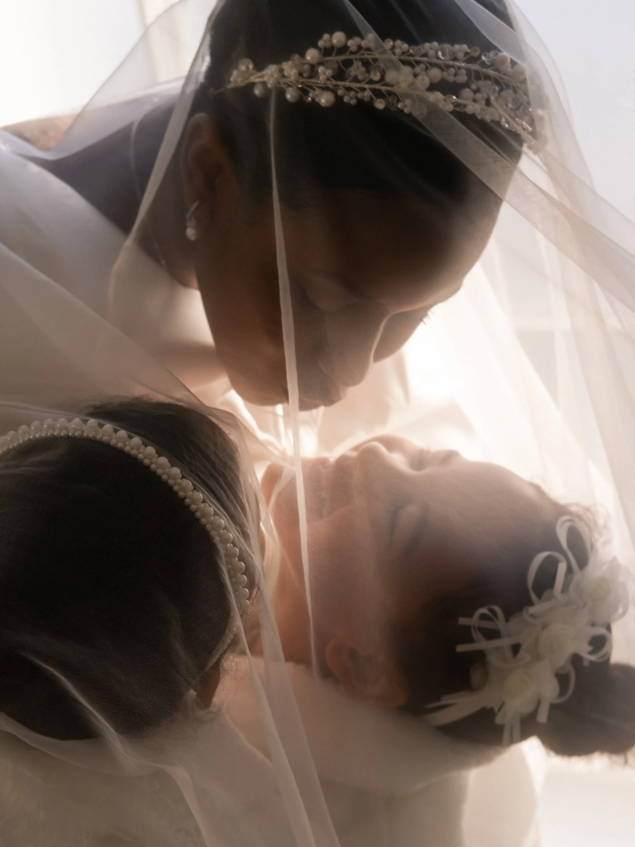 Close-up of a bride and groom kissing behind a wedding veil, with the bride wearing pearl earrings and a pearl-embellished headpiece.