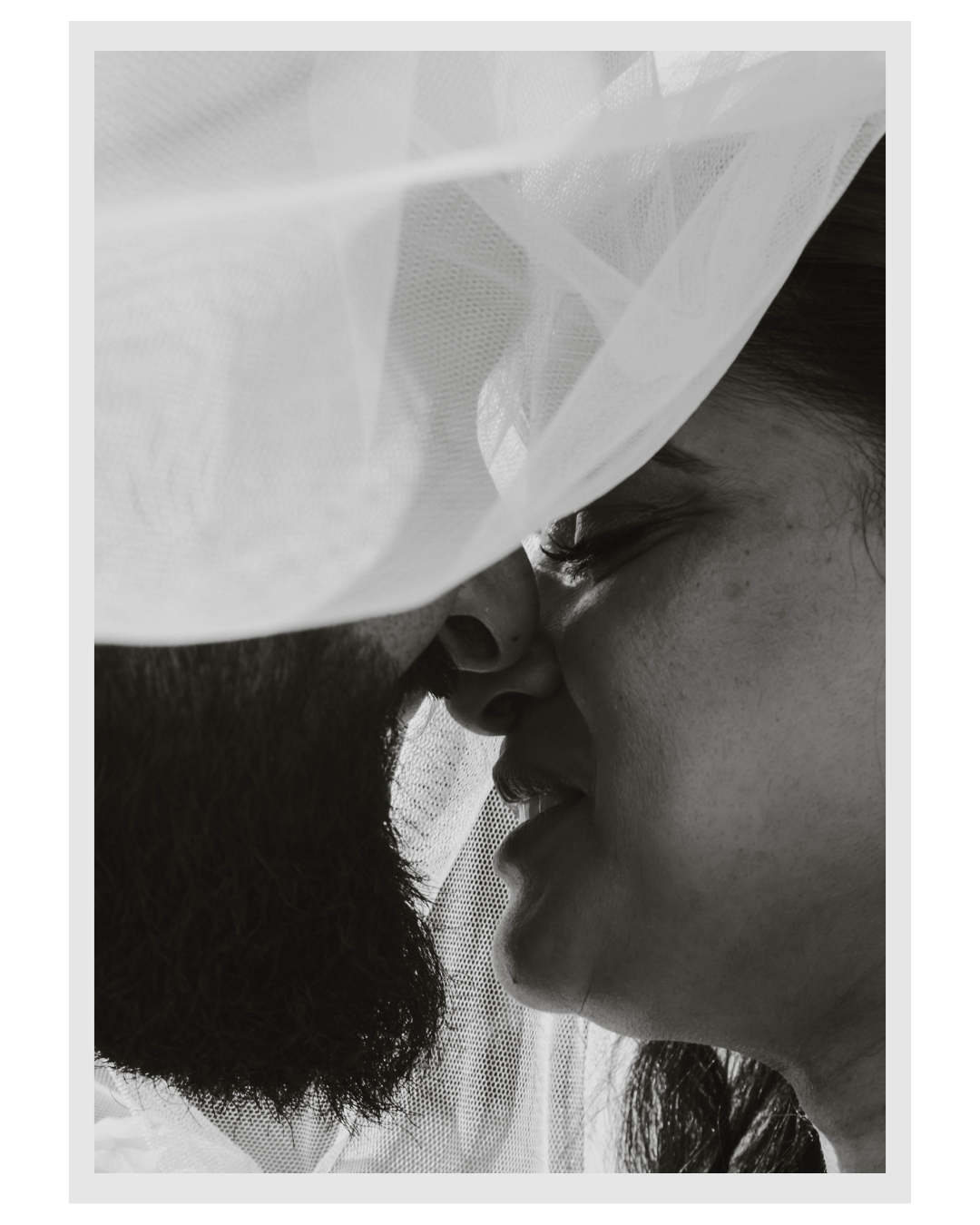 Close-up black and white photo of couple with faces very close, standing under a white veil or fabric, with their noses nearly touching.