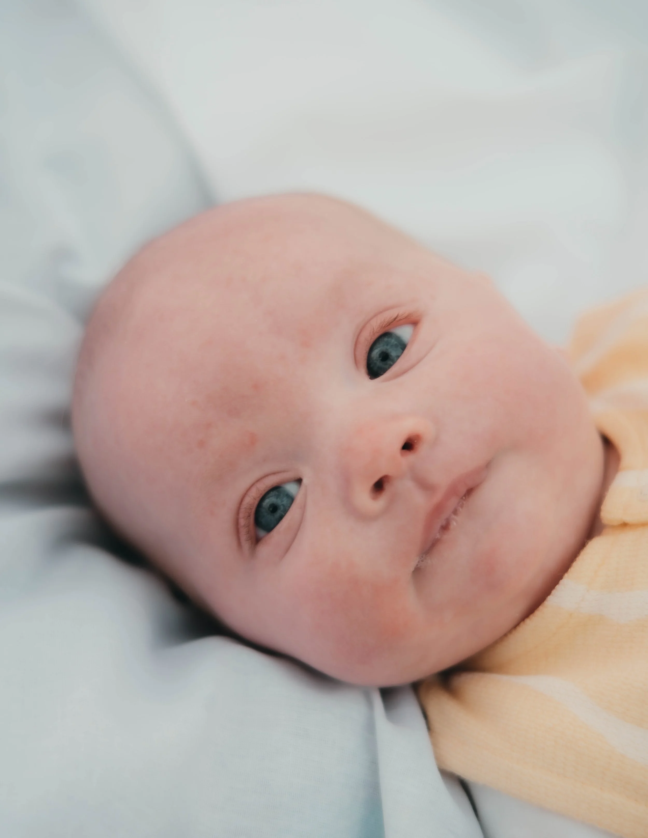 Close-up of a baby's face with blue eyes, lying on a white surface, wearing a light yellow outfit.