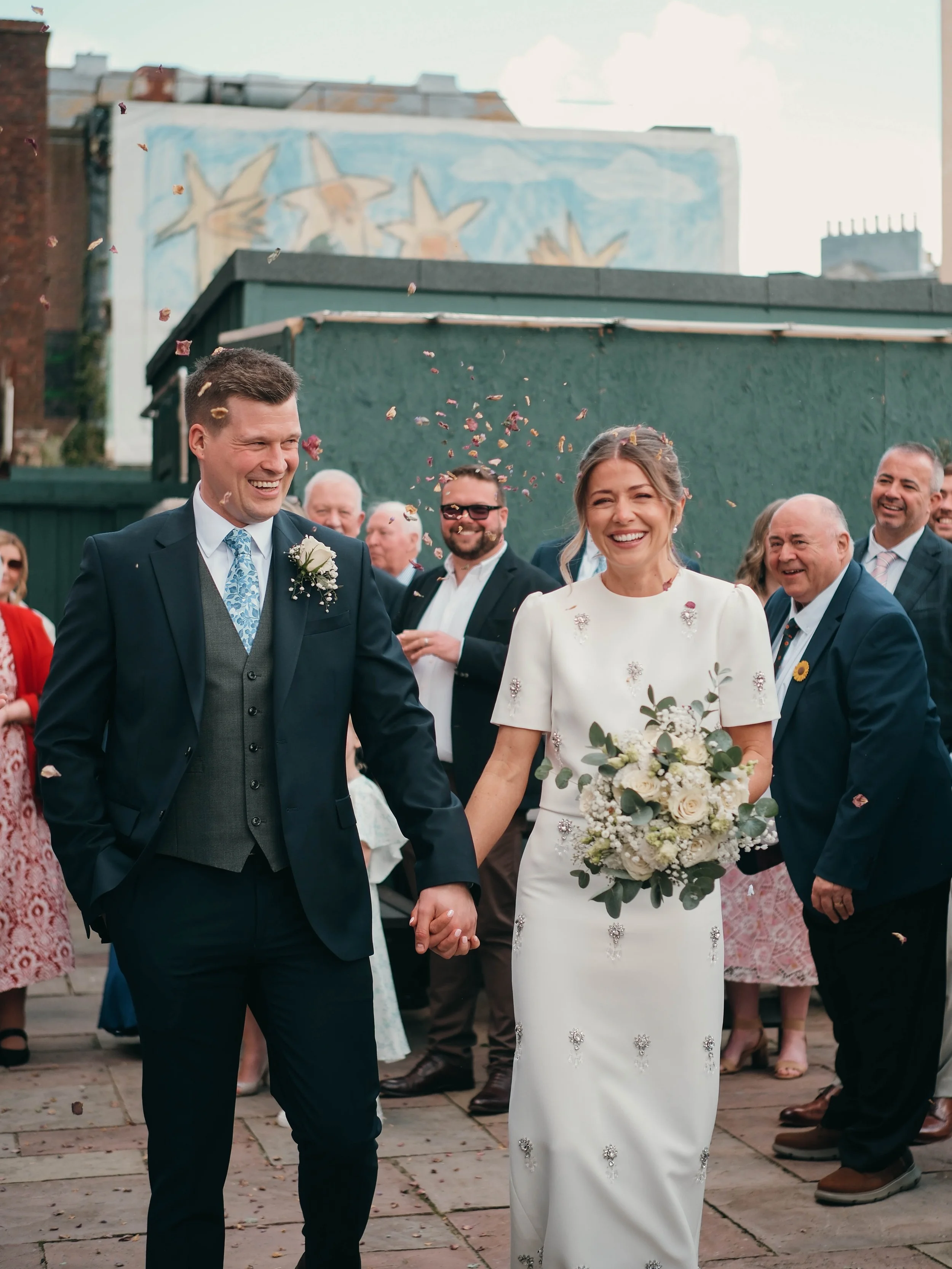 A newlywed couple walking hand in hand outdoors, surrounded by friends and family, with confetti falling around them. The groom wears a black suit and tie, and the bride wears a white wedding dress holding a bouquet. Everyone appears happy and celebr