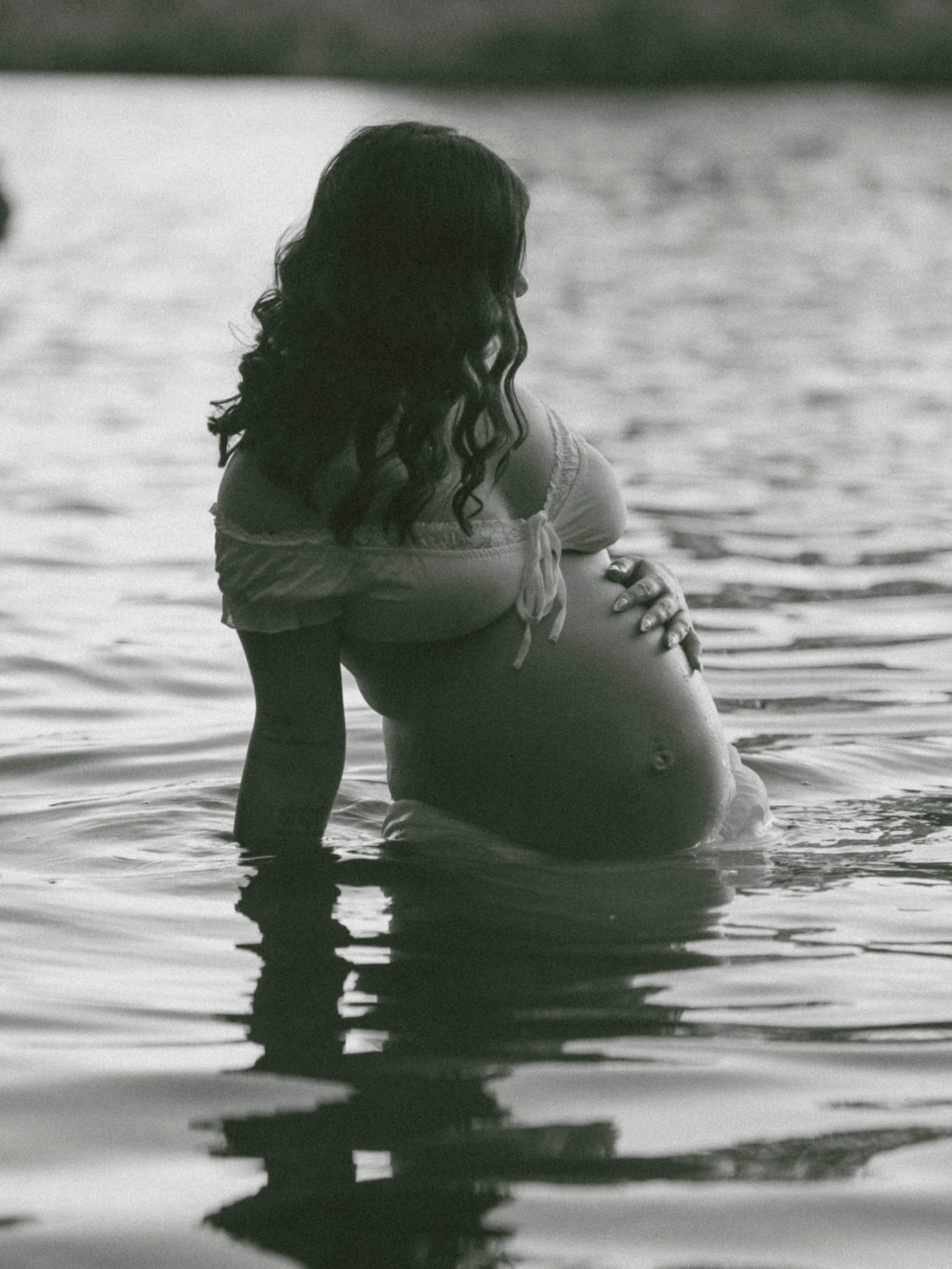 A woman with dark curly hair sitting in water at the edge of a body of water, looking away from the camera.