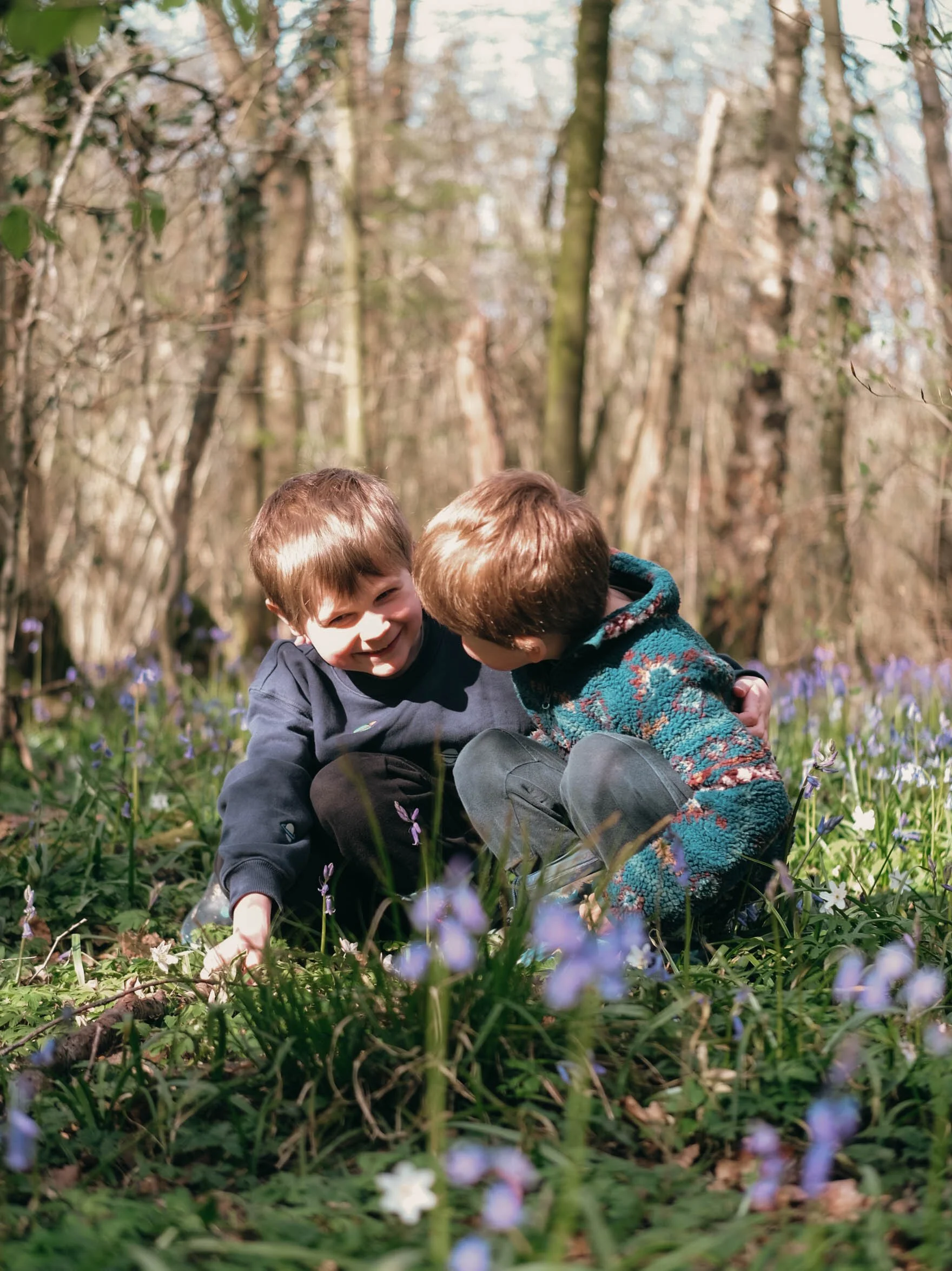 Two young boys are playing in a forested area with wildflowers, smiling and embracing each other.