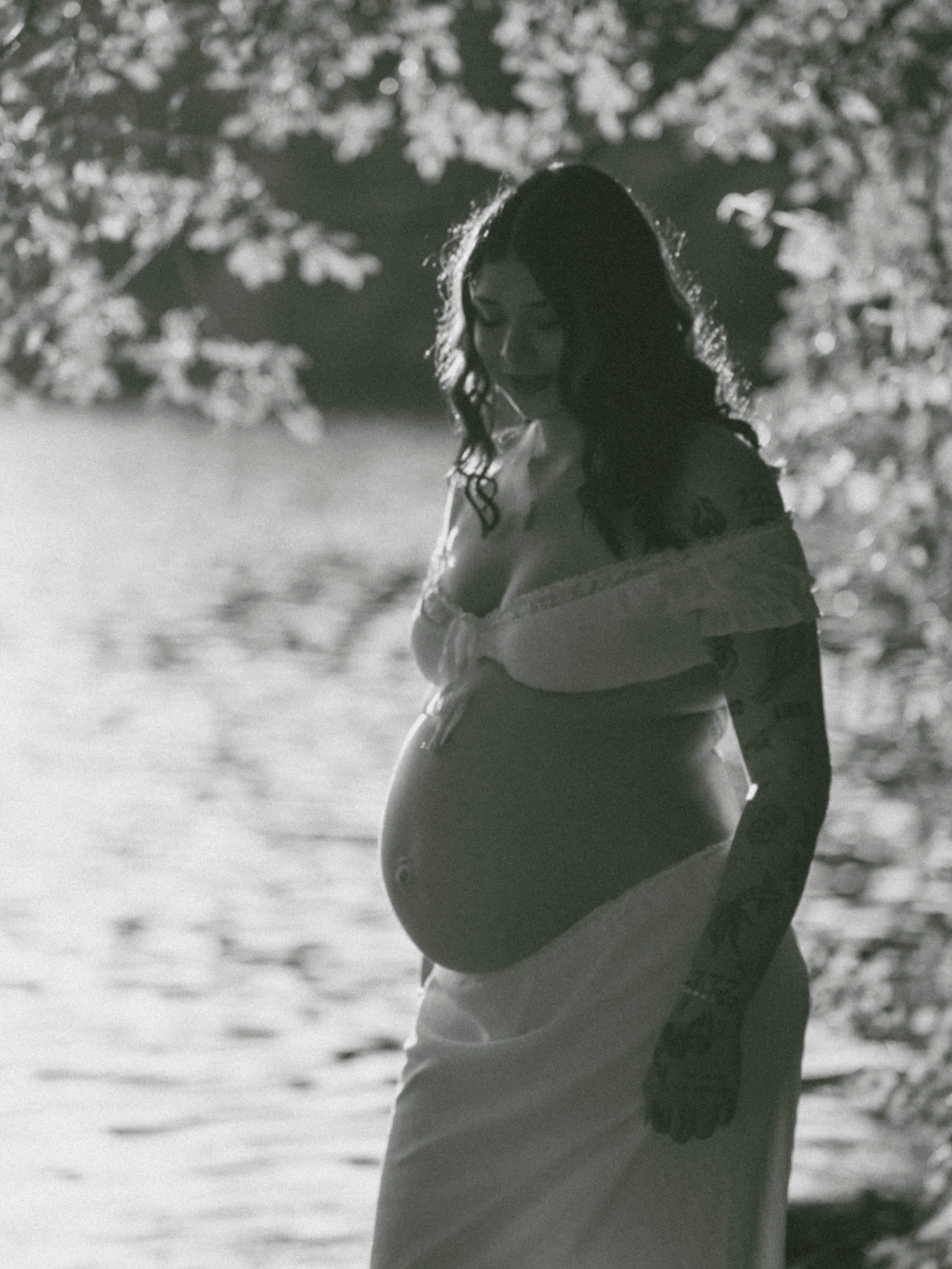 A pregnant woman standing outdoors, cradling her belly, with trees in the background, in a black-and-white photo.