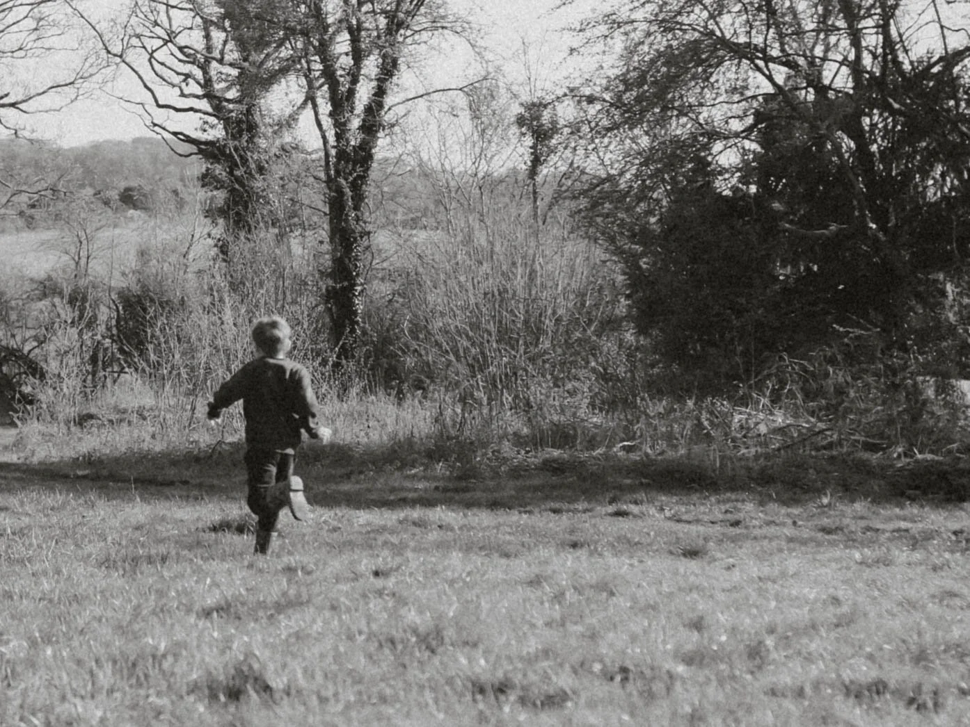 A child running outdoors in a grassy field with trees and bushes in the background, in black and white.