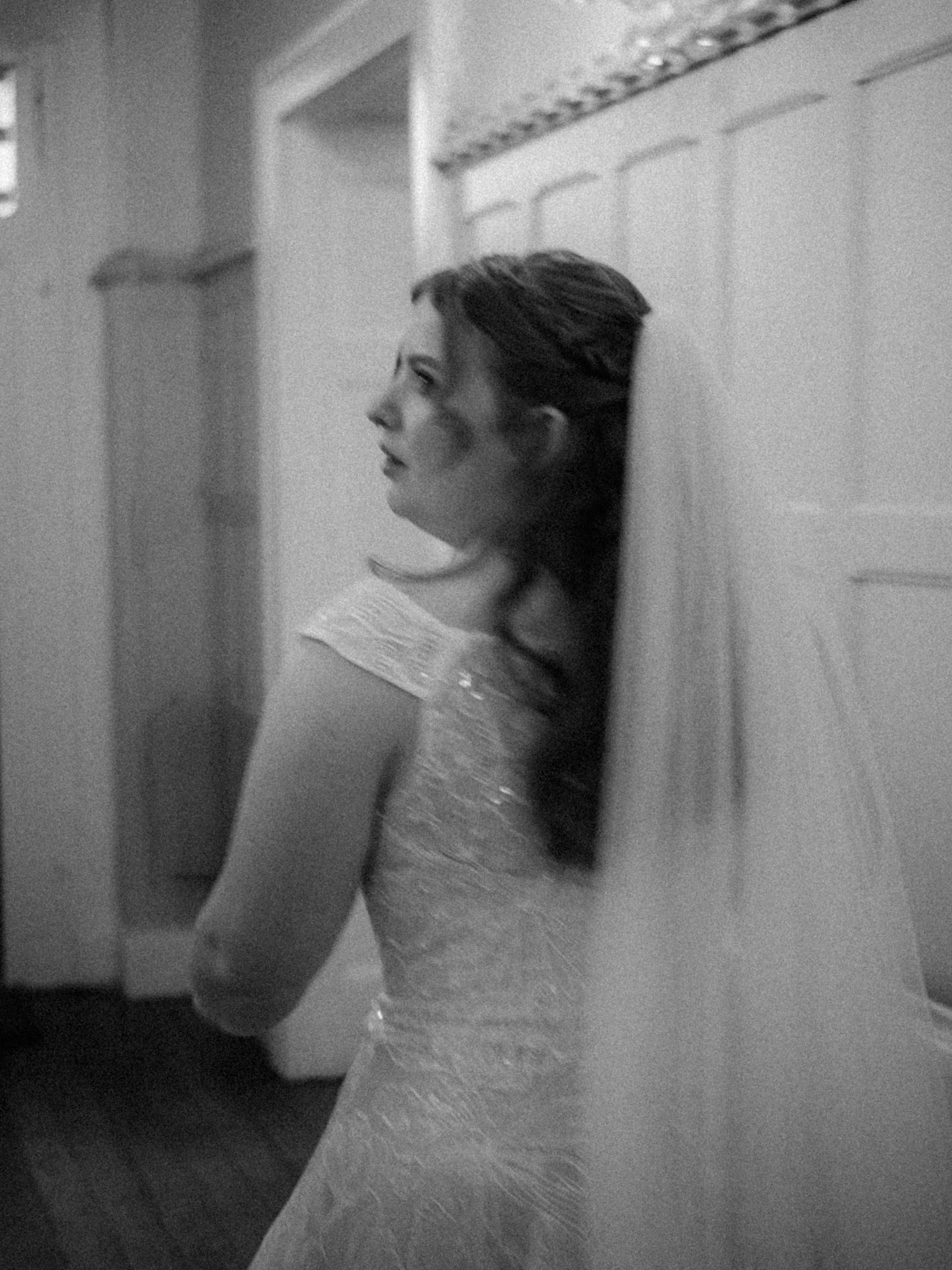 A woman with dark hair styled in an updo, wearing a textured dress, leaning against a wall with decorative molding in a dimly lit interior setting.