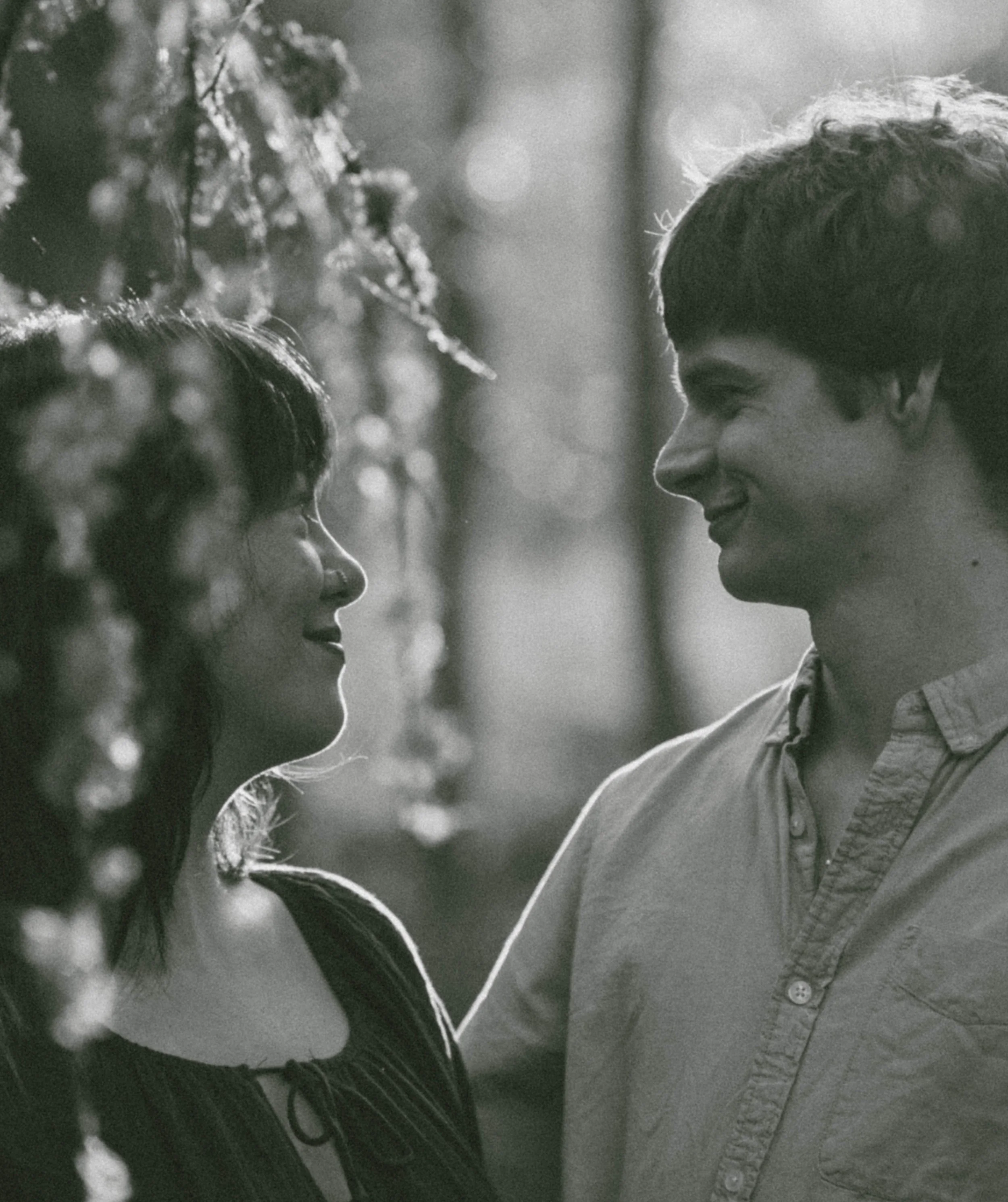 A black-and-white photo of a young boy and woman smiling at each other in a natural outdoor setting, with bokeh lights in the background.