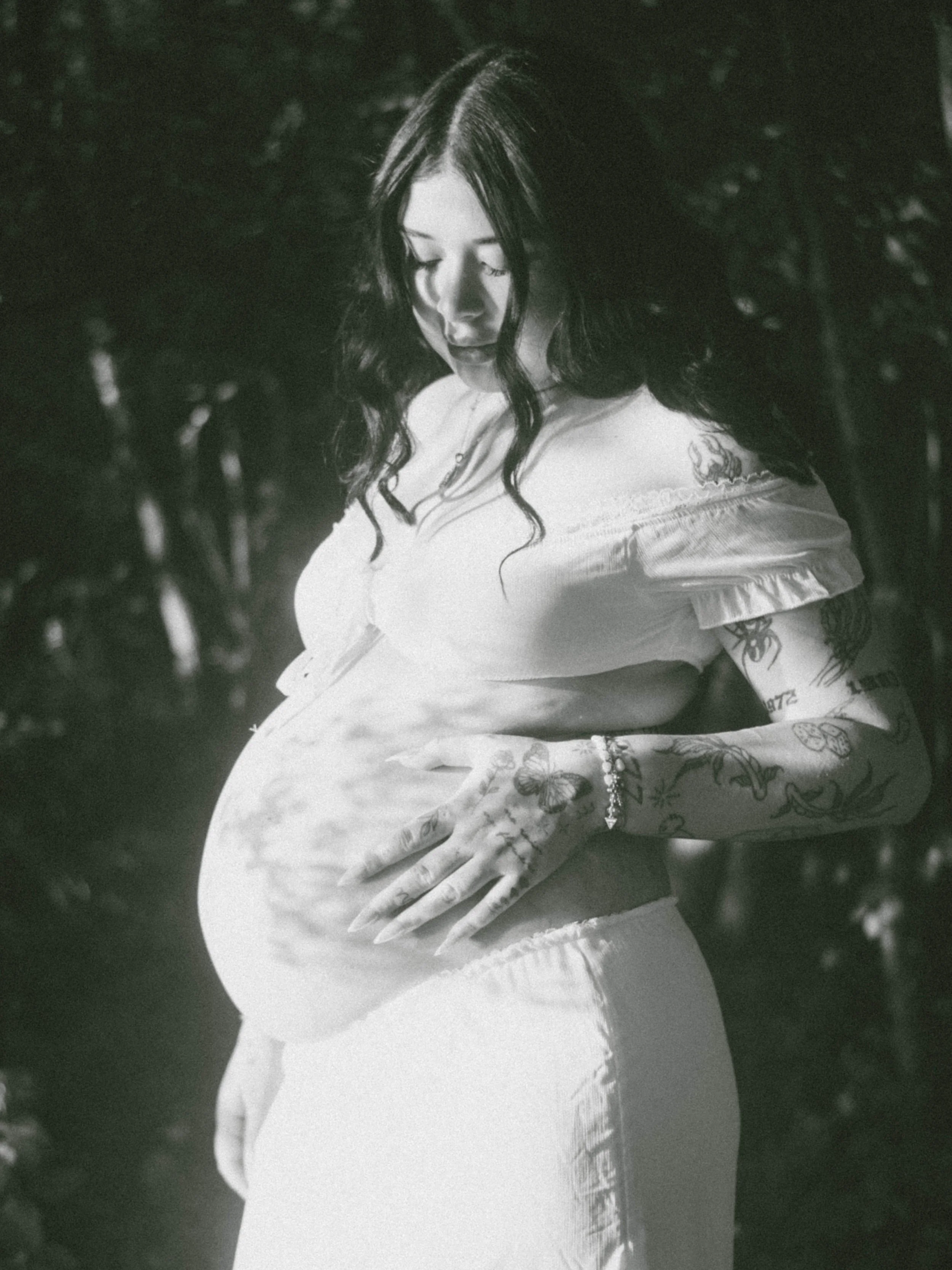 A black and white photo of a woman with long wavy hair, wearing a light-colored off-shoulder top, with tattoos on her arm, looking down with her eyes closed.