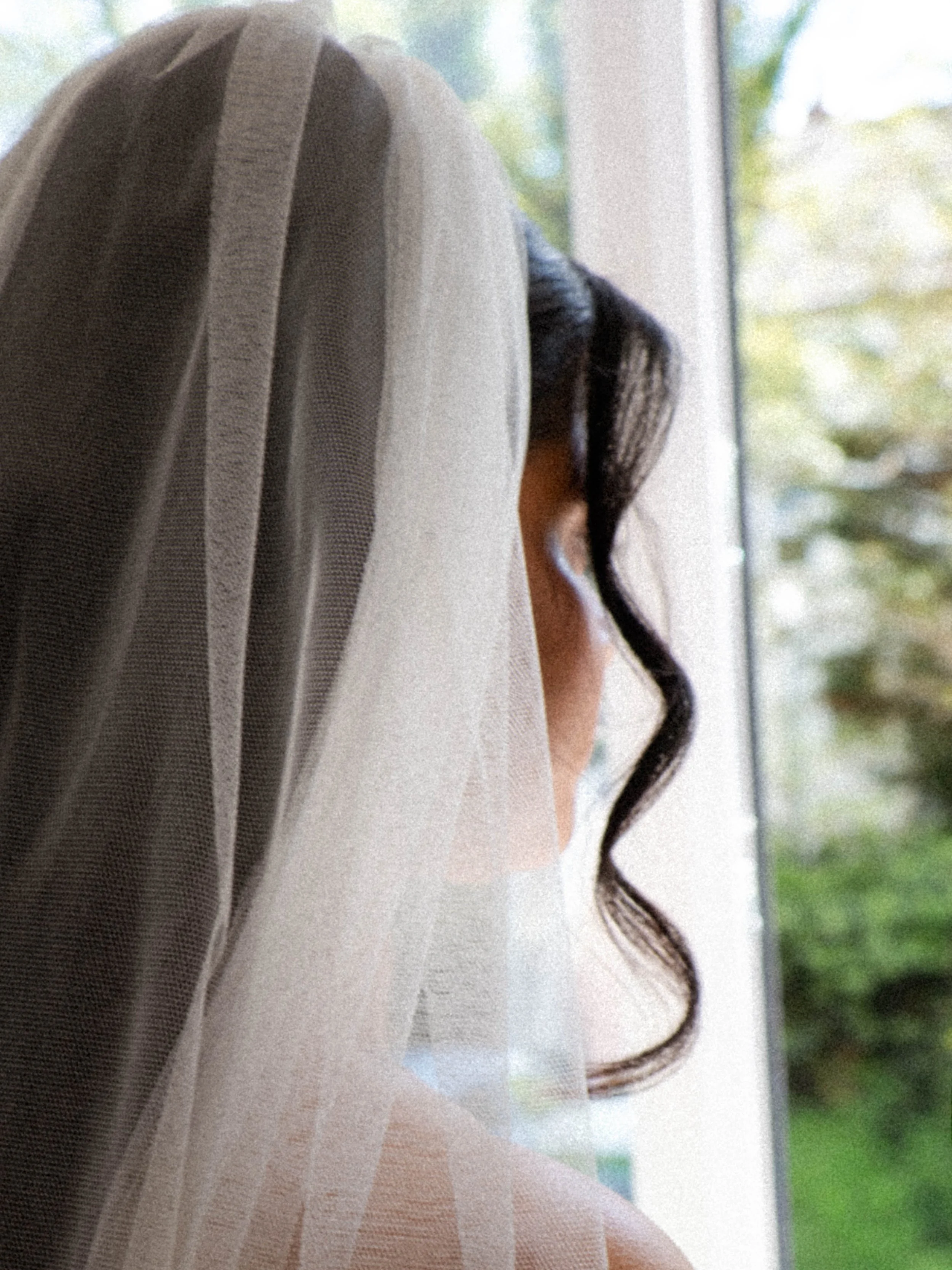 Close-up of a person with long dark hair standing by a window, with greenery outside.