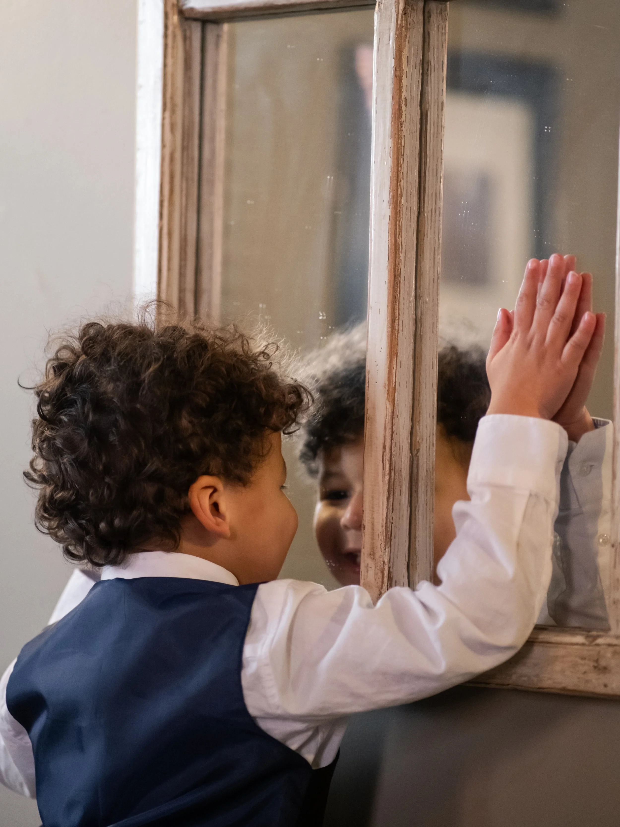 A young boy with curly hair and a dark blue vest over a white shirt is looking at himself in an old-fashioned mirror with a wooden frame. The boy is touching the mirror with his right hand, and his reflection can be seen in the mirror.