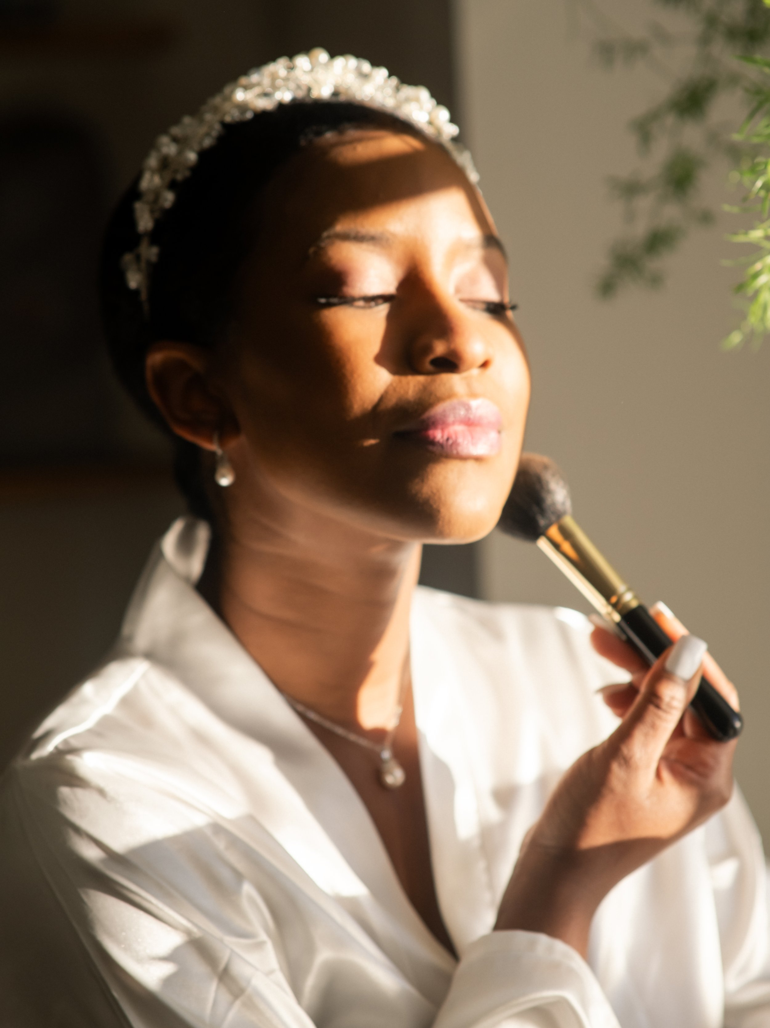 A woman with closed eyes, wearing a tiara and earrings, applying makeup with a brush, dressed in a white silk robe, in natural sunlight.