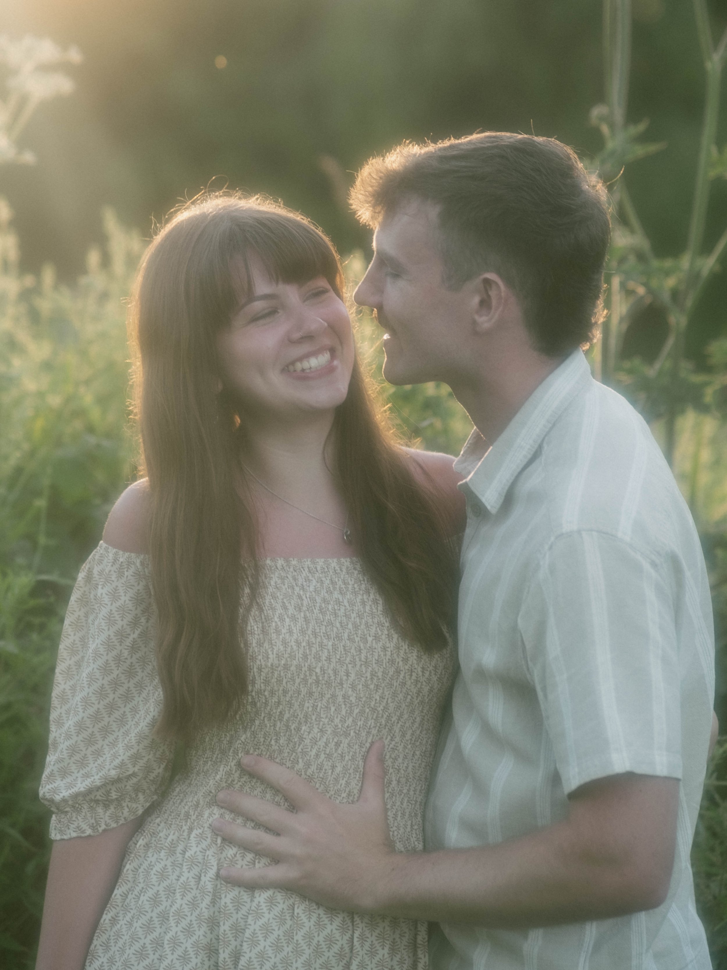 A young woman with long brown hair smiling as a young man with short brown hair leans in close, outdoors during golden hour, greenery in the background.