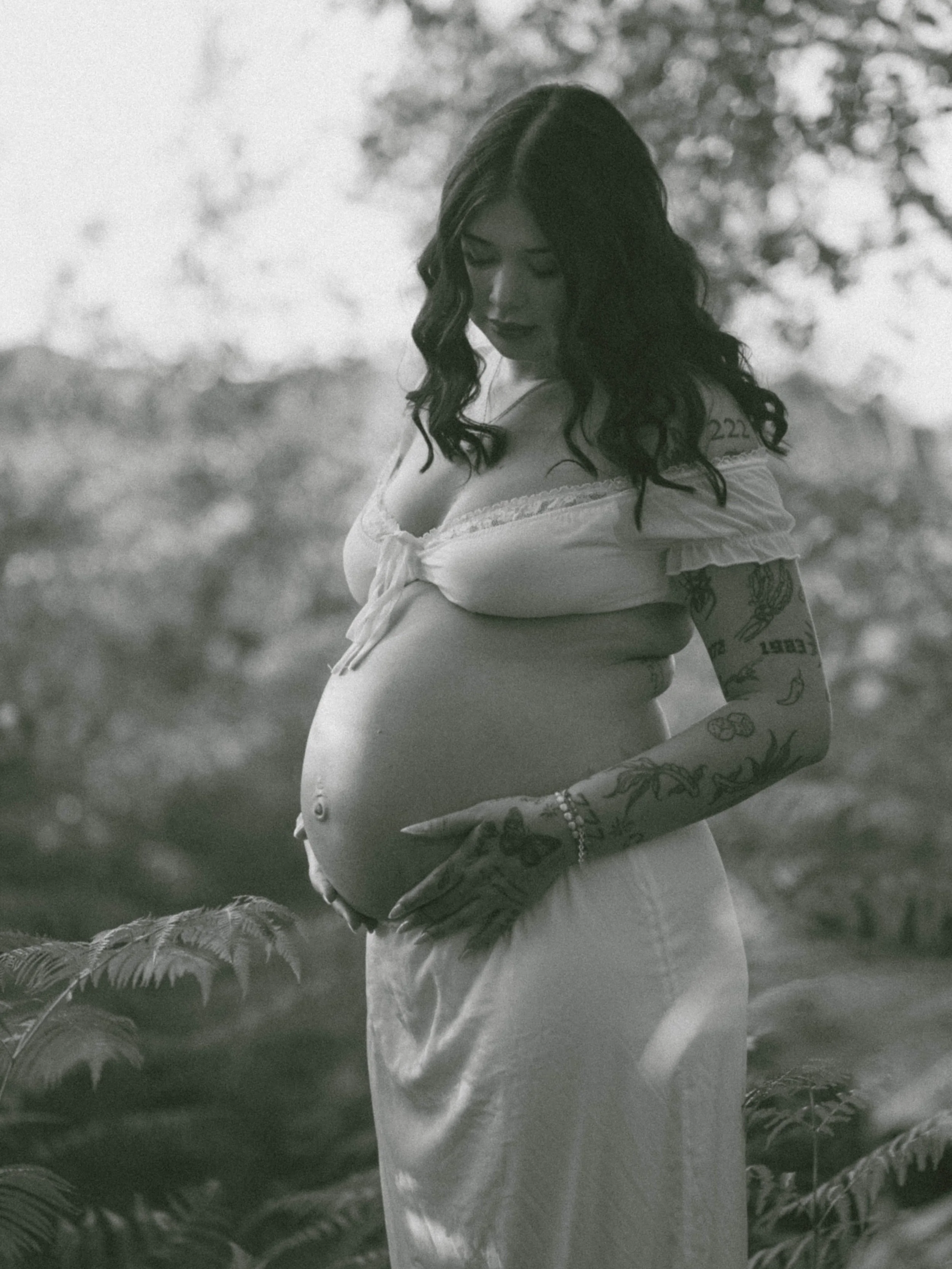 A pregnant woman with dark, curly hair and tattoos on her arms and shoulder, standing outdoors in a natural setting with blurred trees and foliage in the background, wearing a light-colored off-the-shoulder top and a long skirt.