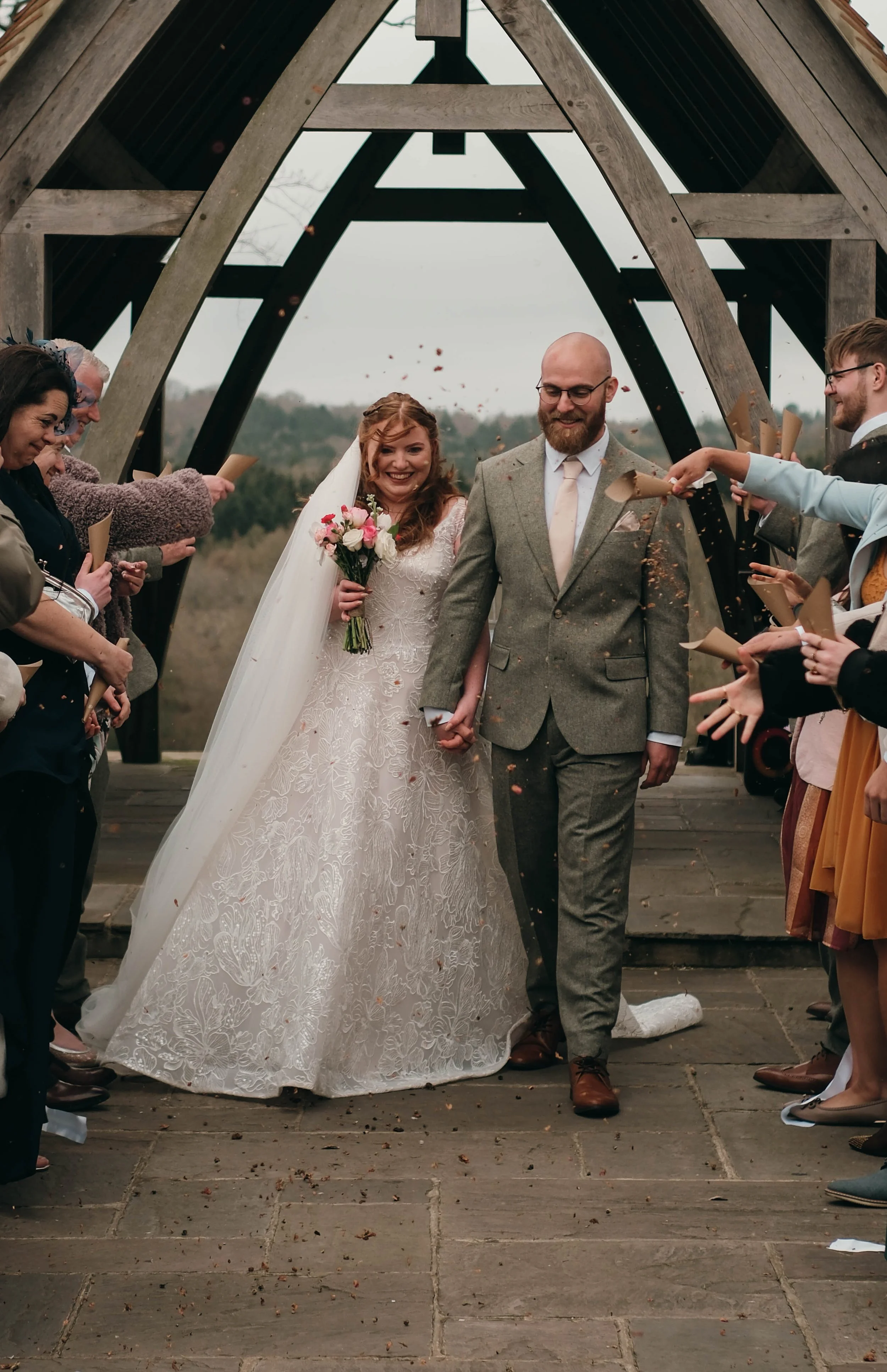 A bride and groom walking hand in hand during their wedding celebration, surrounded by guests throwing confetti beneath a wooden A-frame structure outdoors.