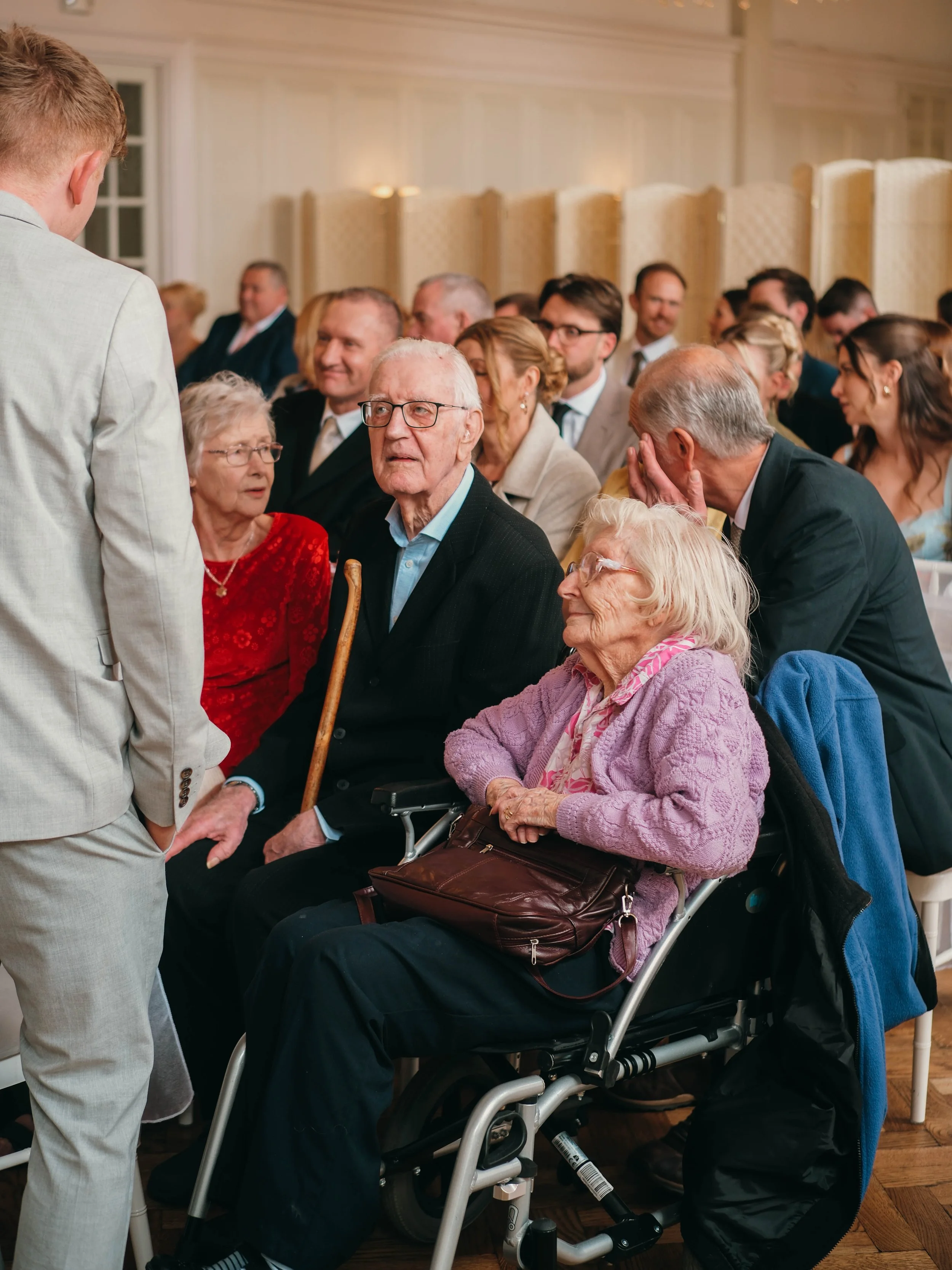 A man in a beige suit is talking to a group of elderly people, including a woman in a red dress and a man in glasses seated in a wheelchair, at a social gathering in a well-lit room with wooden floors and cream-colored room dividers.