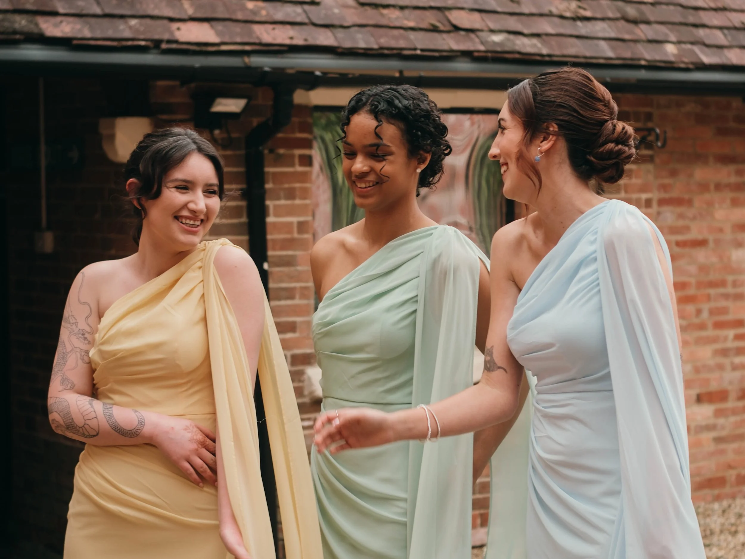 Three women dressed in pastel-colored dresses are standing outdoors in front of a brick house with a shingled roof, engaged in conversation and smiling.