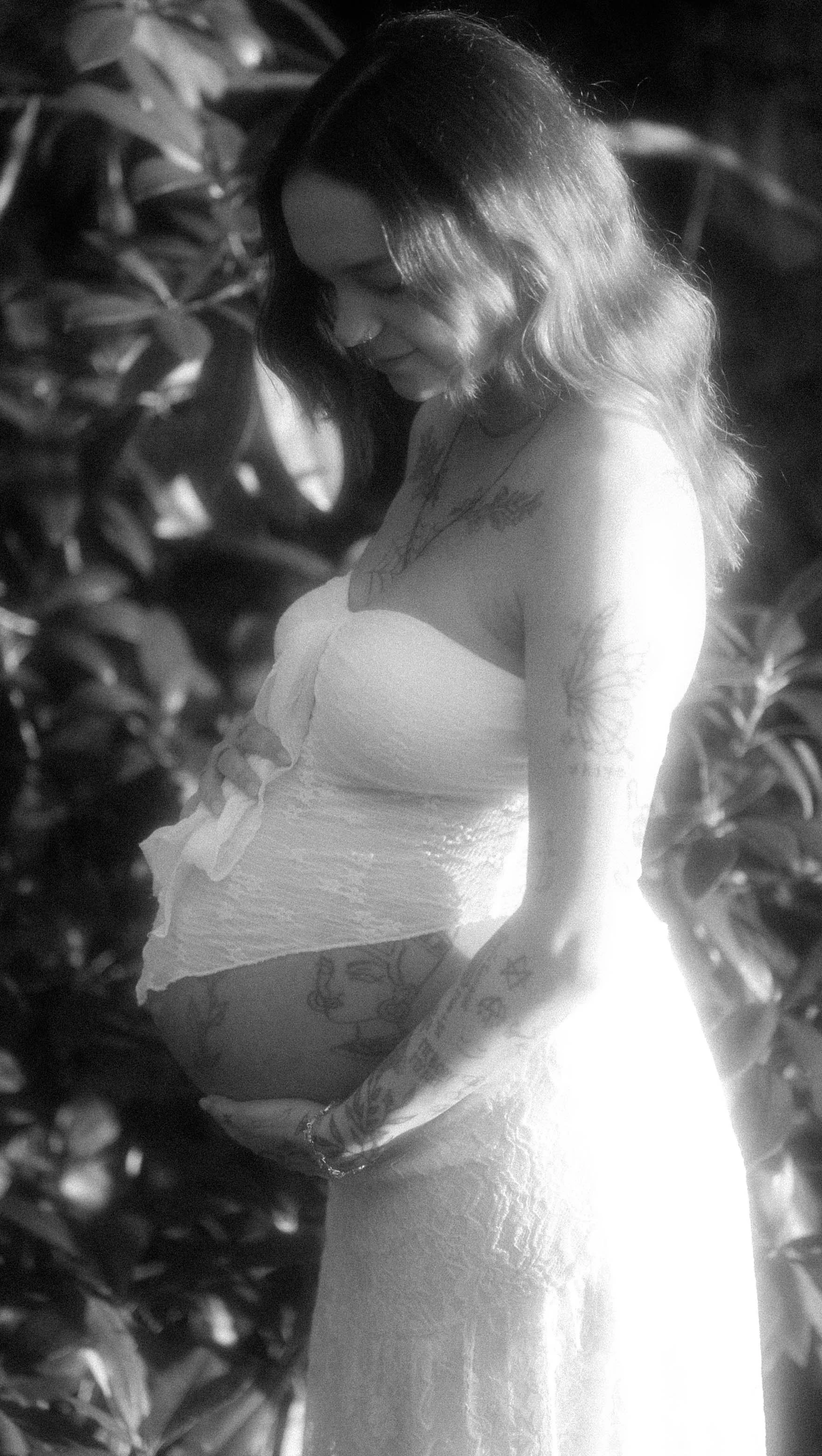 A woman with tattoos on her arm and chest, wearing a strapless top, posed outdoors beside foliage, in black and white.