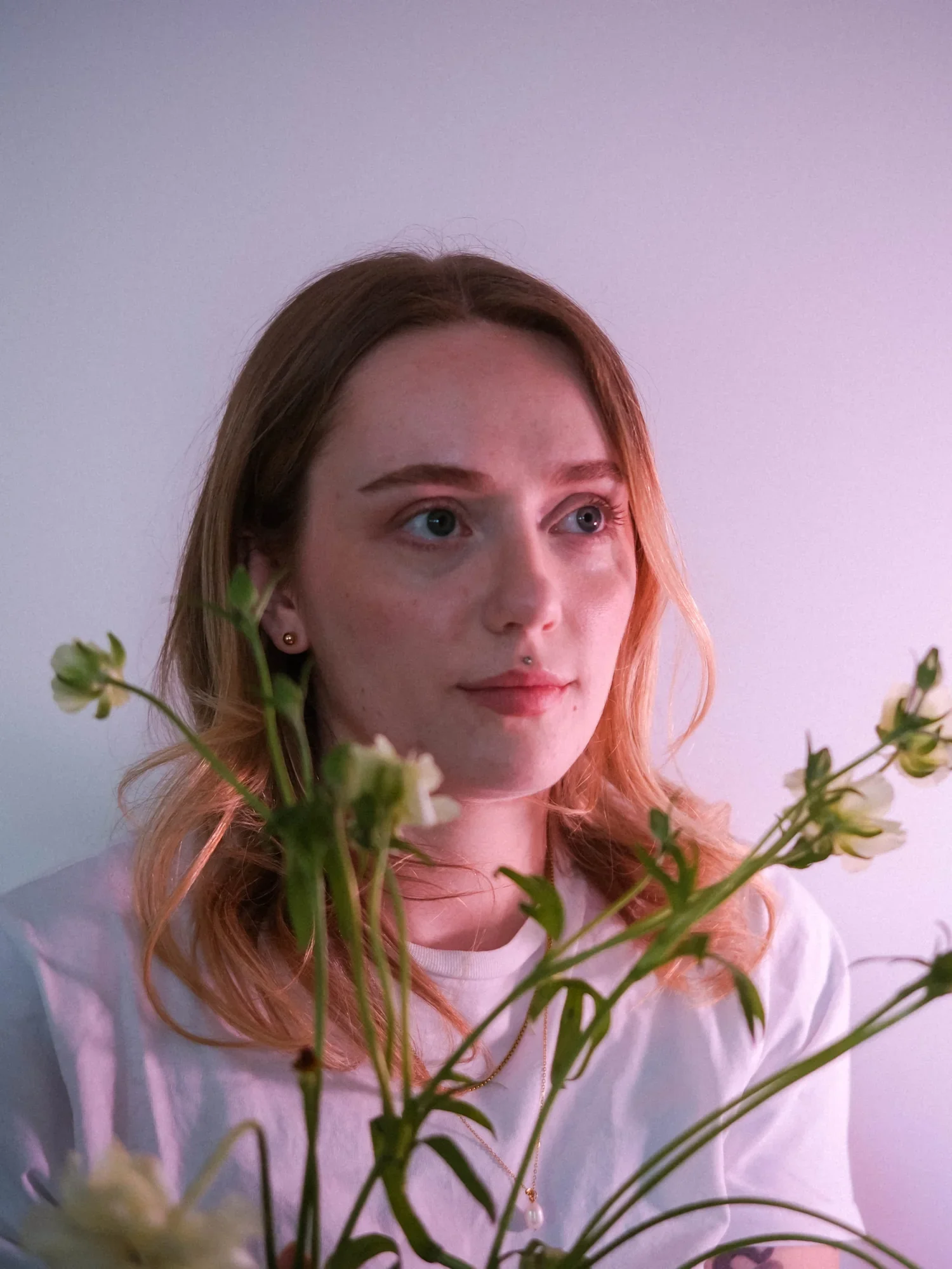 A woman with red hair and light skin looking to the side, surrounded by flowers, indoors with a plain, pastel-colored wall in the background.