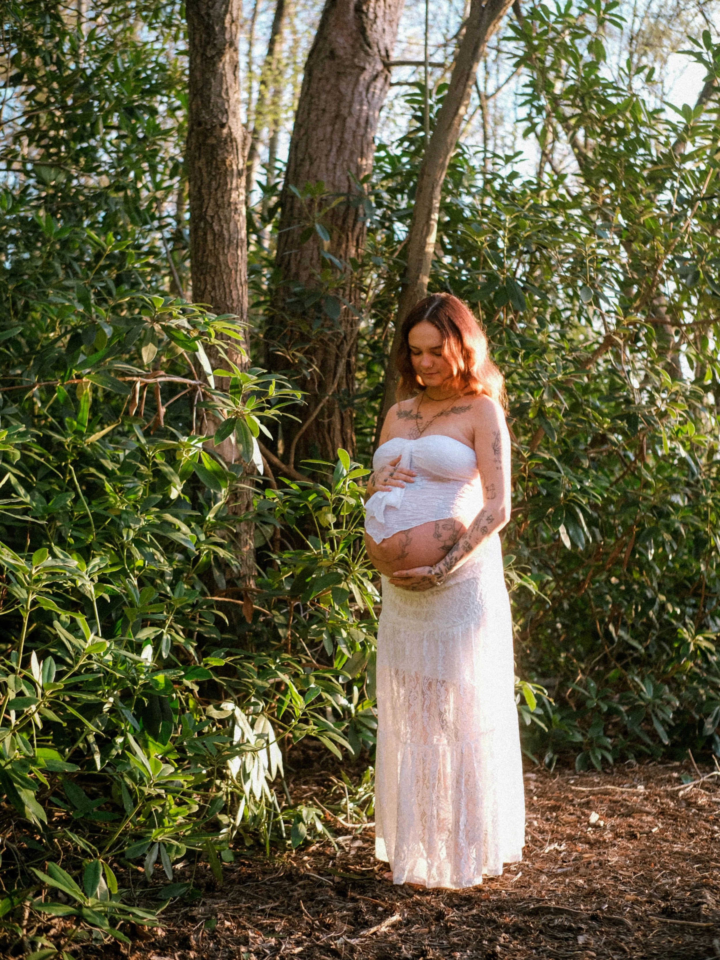 A woman with tattoos is standing outdoors in a wooded area, wearing a white lace dress, with her eyes closed and her hands on her pregnant belly.