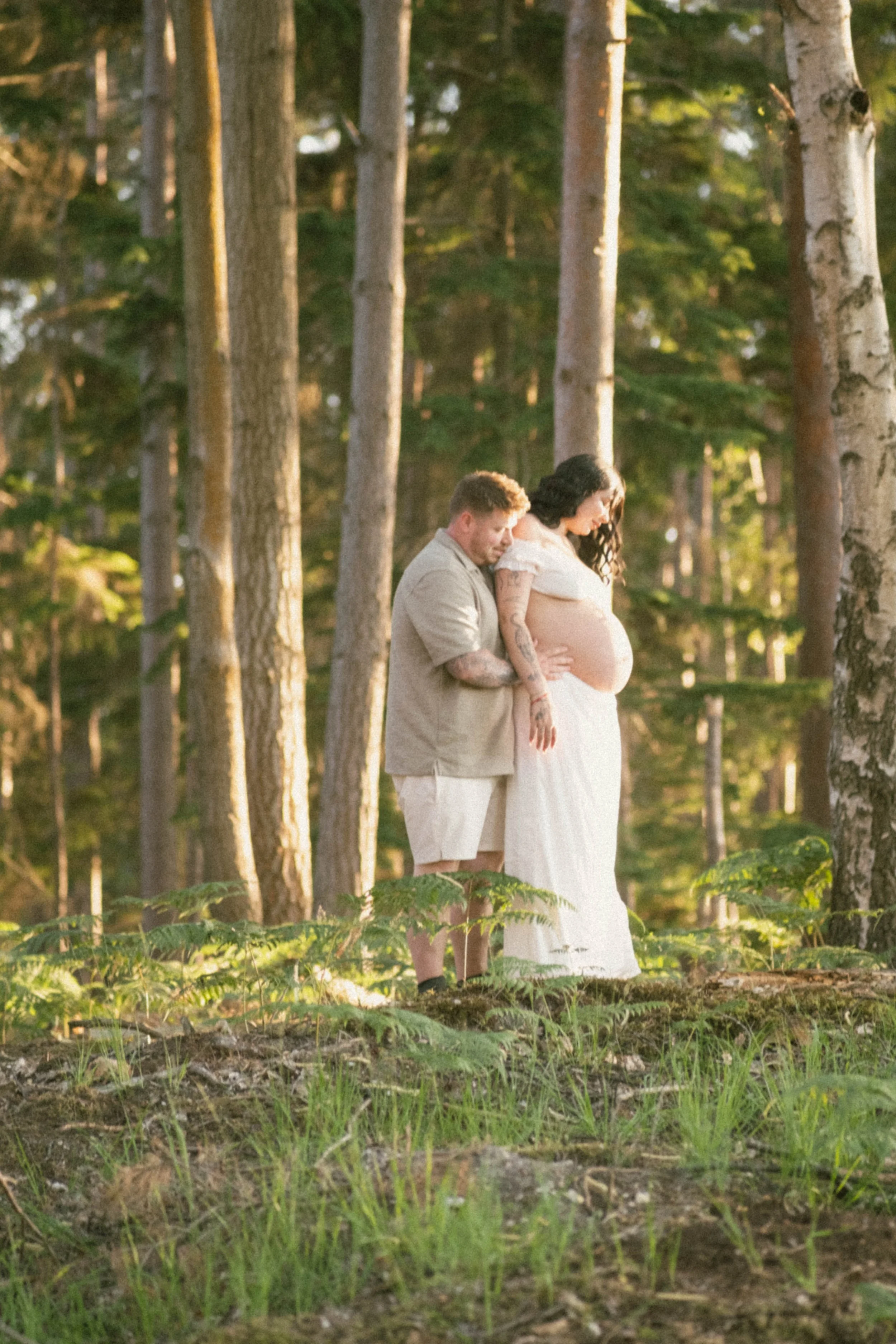 A couple is standing in a forest, with the man behind the pregnant woman, both holding her belly. The woman is wearing a white dress and has dark, wavy hair; the man is dressed in beige with short hair. The scene is lit with warm sunlight, and trees 
