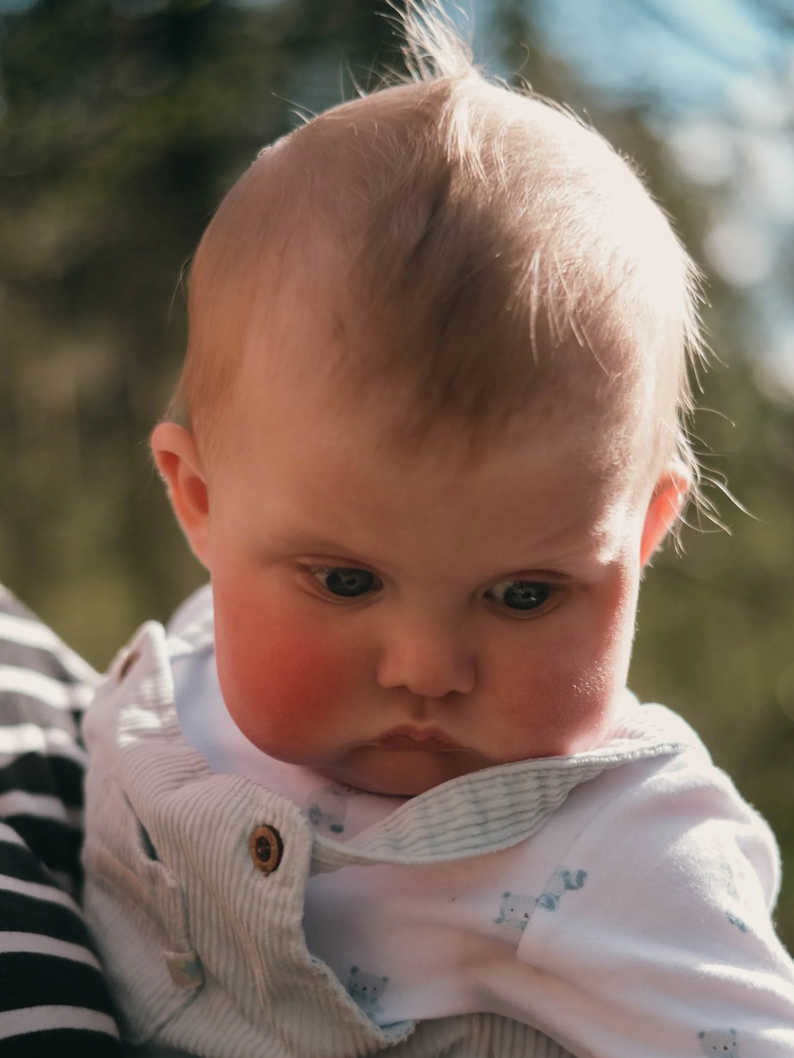 Close-up of a young child's face with blond hair and blue eyes, wearing a white shirt and a light-colored vest with buttons, outdoors with blurred greenery in the background.