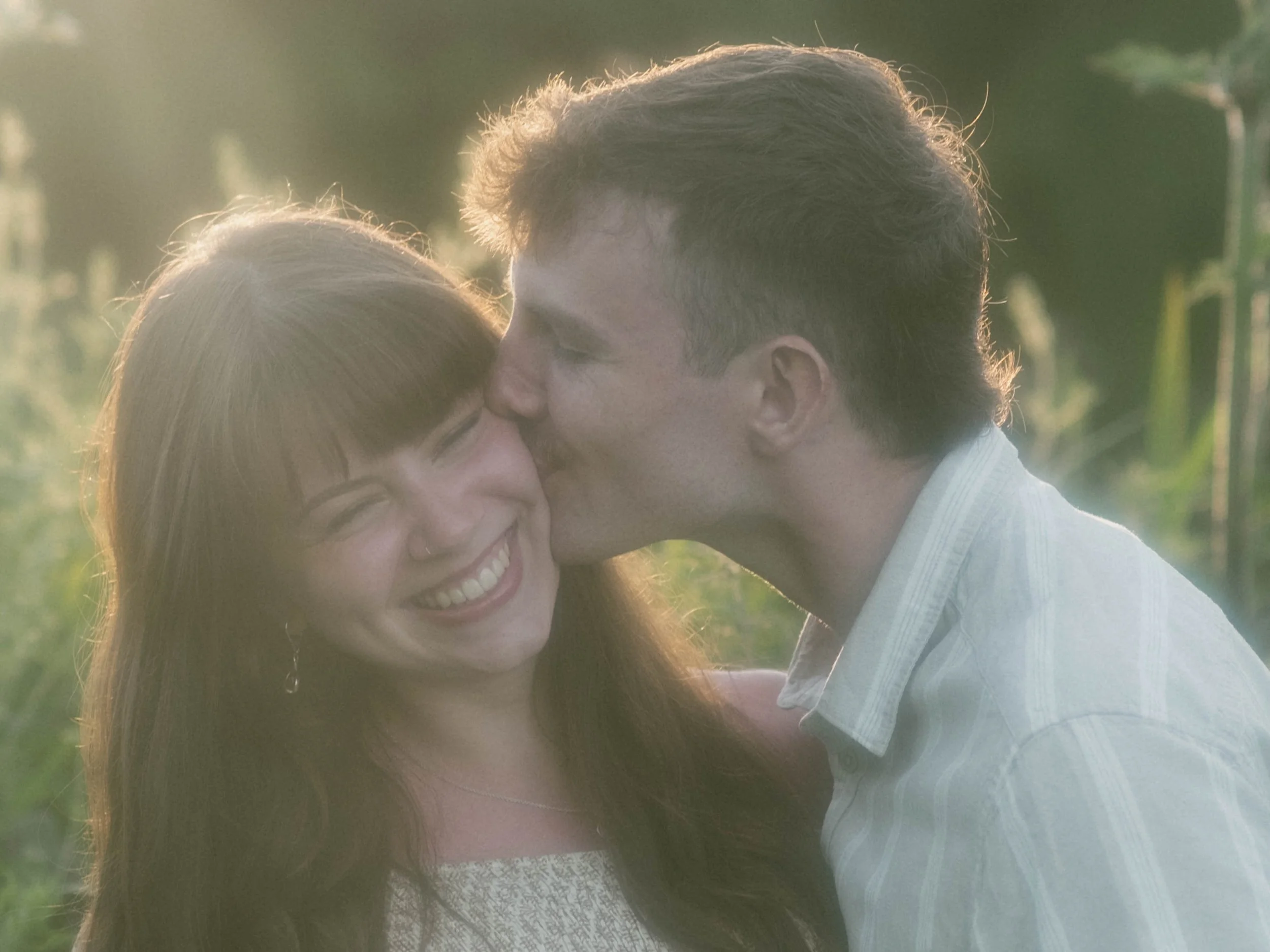 A couple embracing outdoors during golden hour, with the man kissing the woman on the forehead, both smiling, in a lush green setting.
