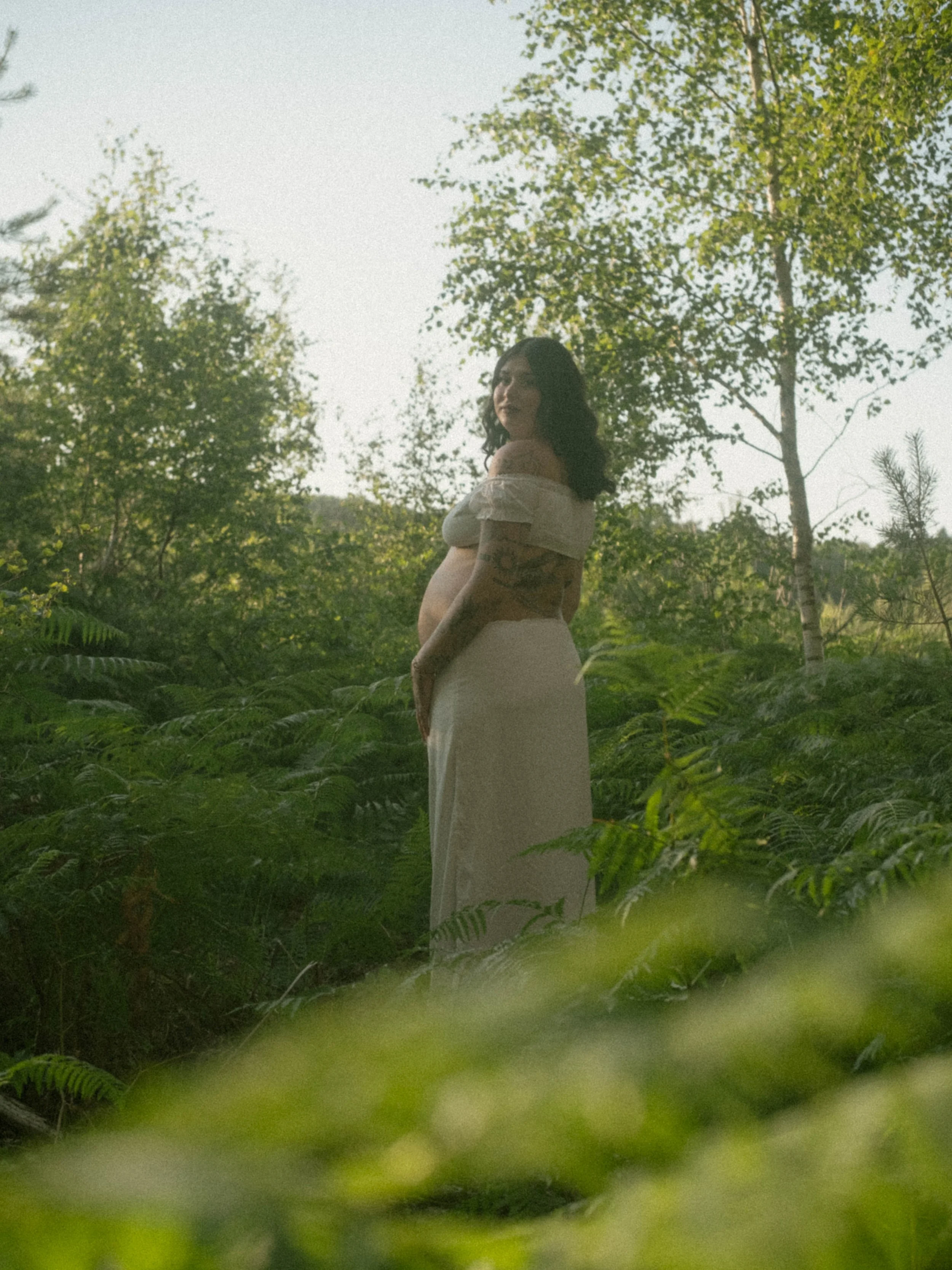 A pregnant woman standing in a lush green outdoor setting, surrounded by trees and foliage, with a serene expression and relaxed pose.
