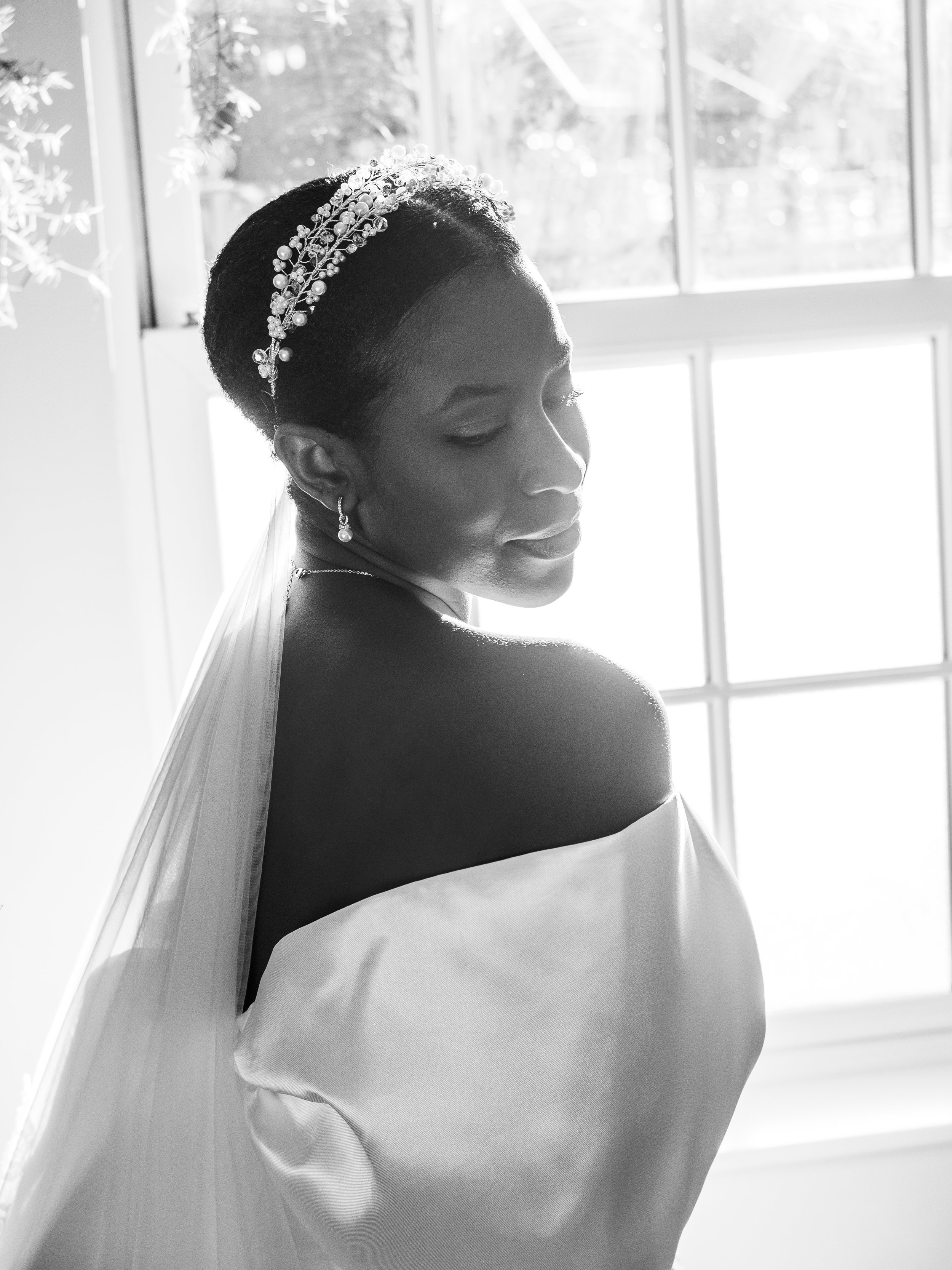 Black and white image of a woman in a wedding dress with an off-shoulder design, wearing a pearl and crystal headband and matching earrings, standing by a window with sunlight streaming in.