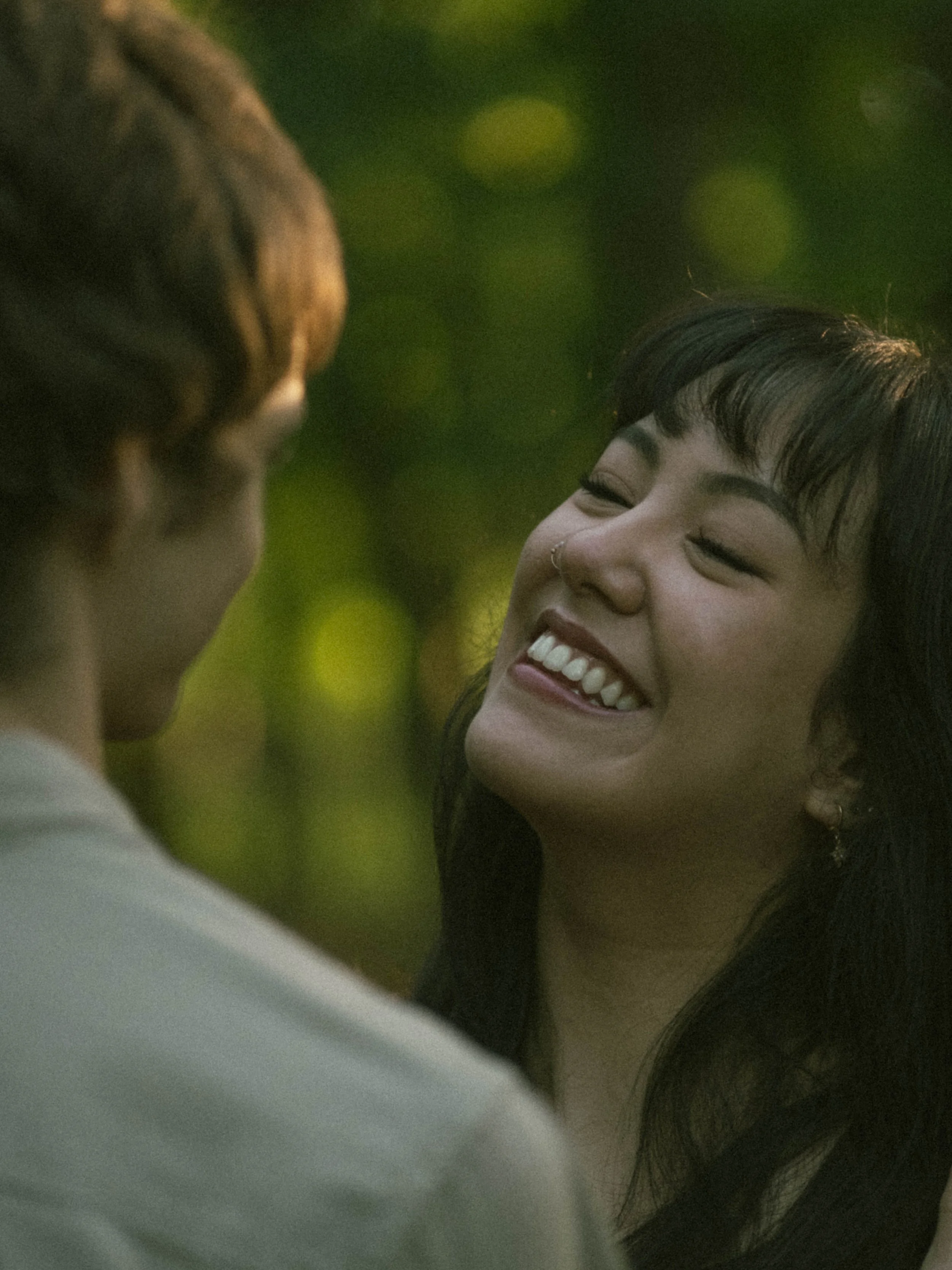 A woman with short black hair and a nose piercing smiling and appearing happy, with another person partially visible beside her, outdoors with blurred green background.