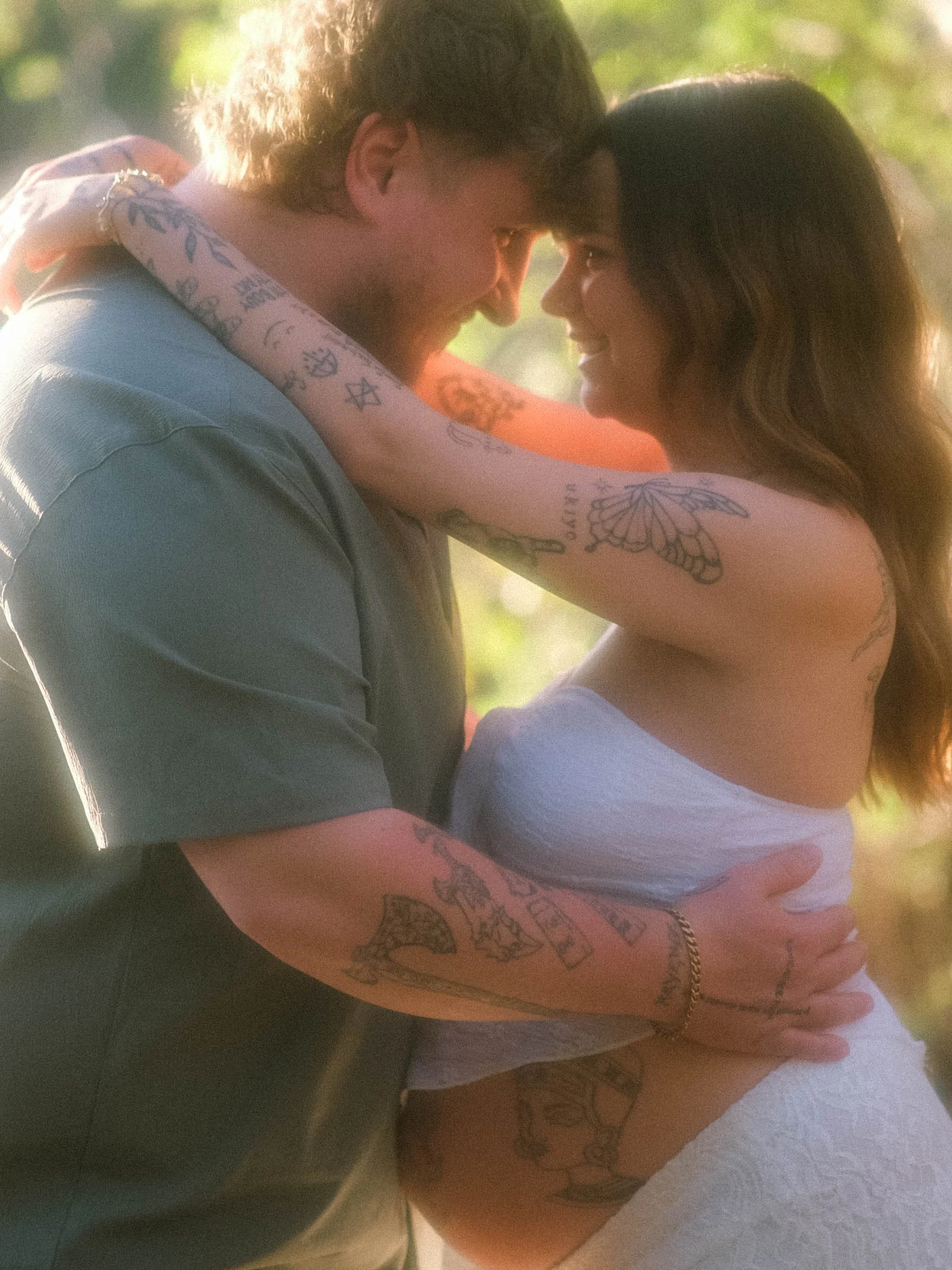 A man and woman are close together, smiling and touching forehead to forehead, with their eyes closed in a tender moment. The man has tattoos on his arms and shoulder, and the woman has long dark hair, with a tattoo on her arm. The background is blur