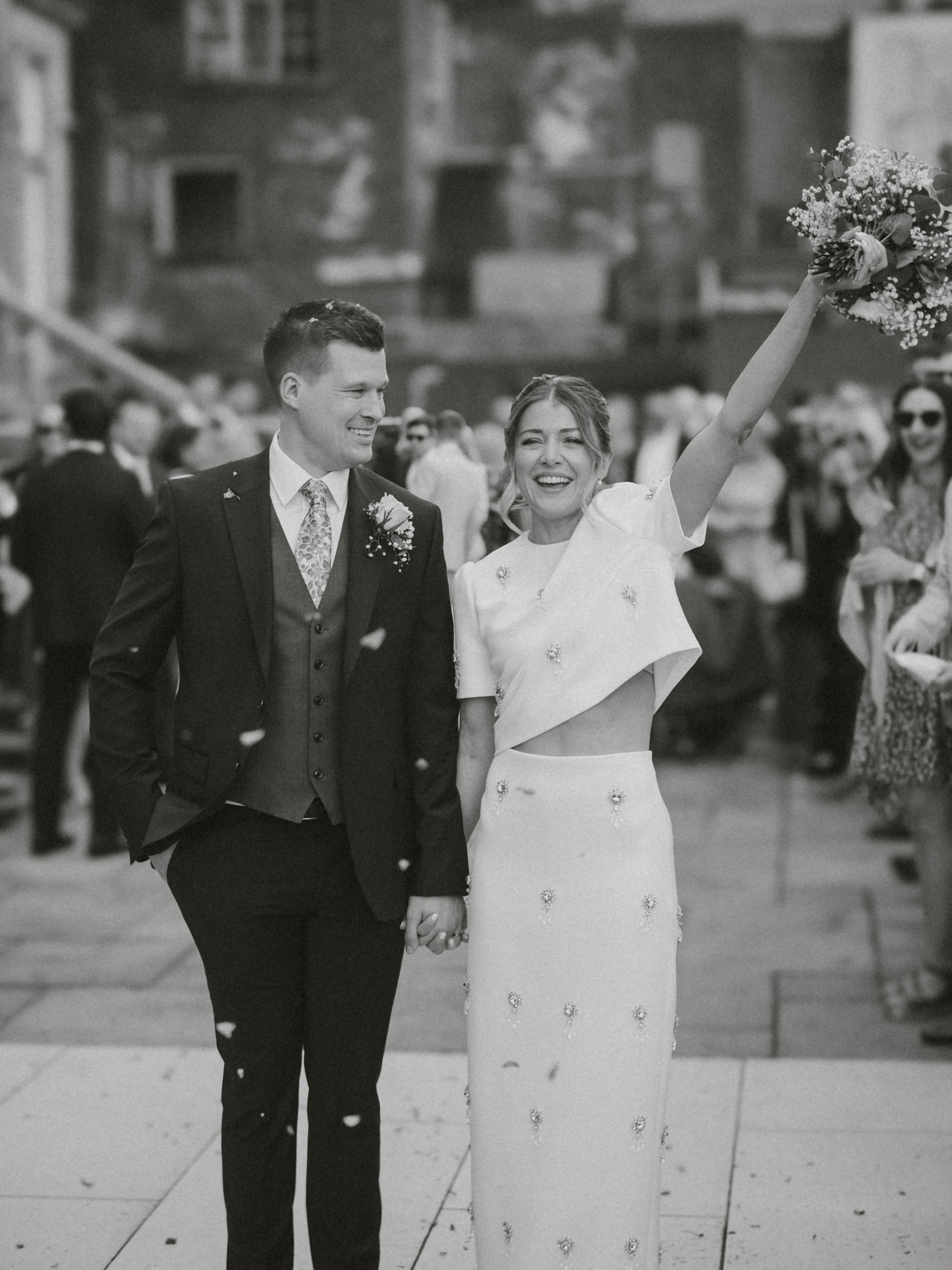 A bride and groom holding hands and smiling, with the bride raising a bouquet in the air, surrounded by guests at their wedding celebration.