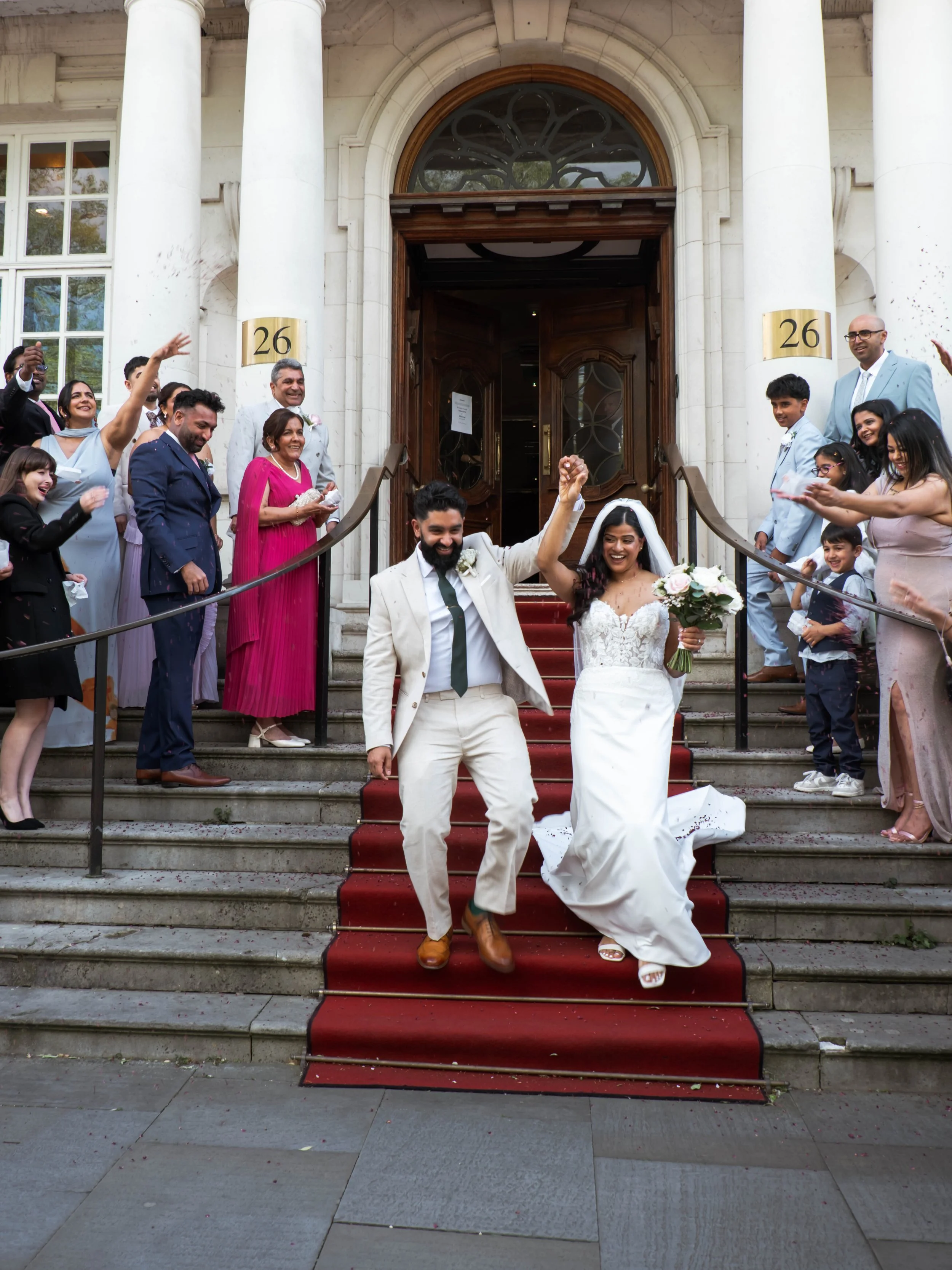 Married couple holding hands and smiling on the stairs outside a church after their wedding, surrounded by family and friends celebrating.