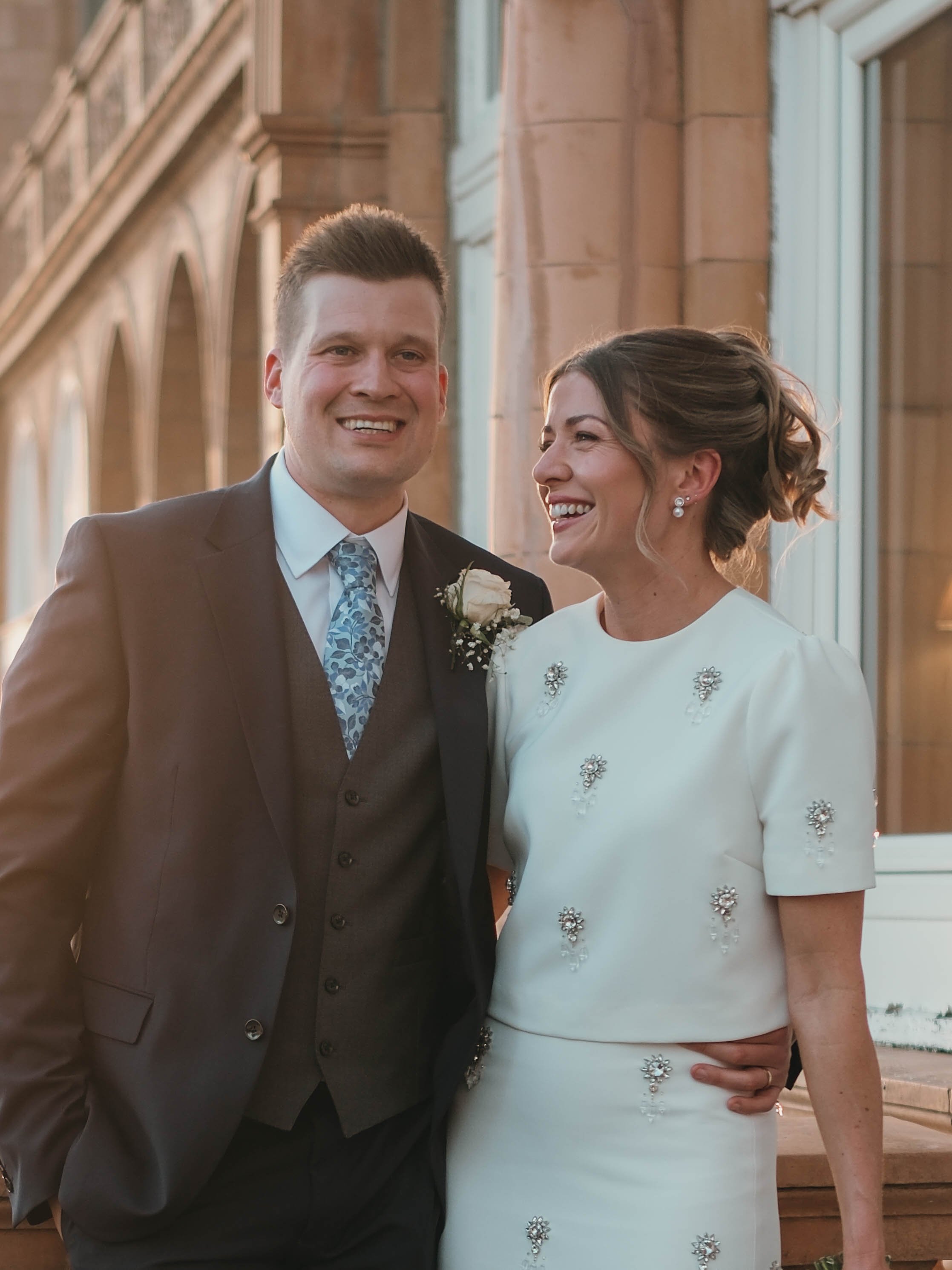 A smiling couple dressed in wedding attire standing close together indoors near large windows with wooden frames.