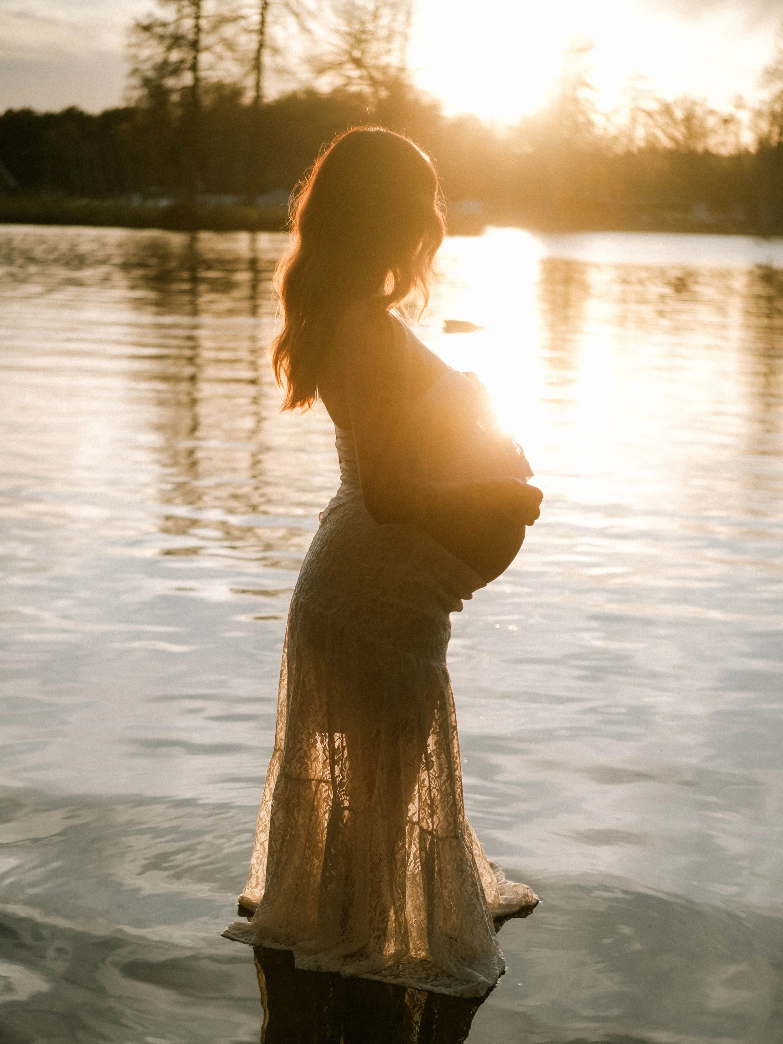 A pregnant woman standing in water during sunset.