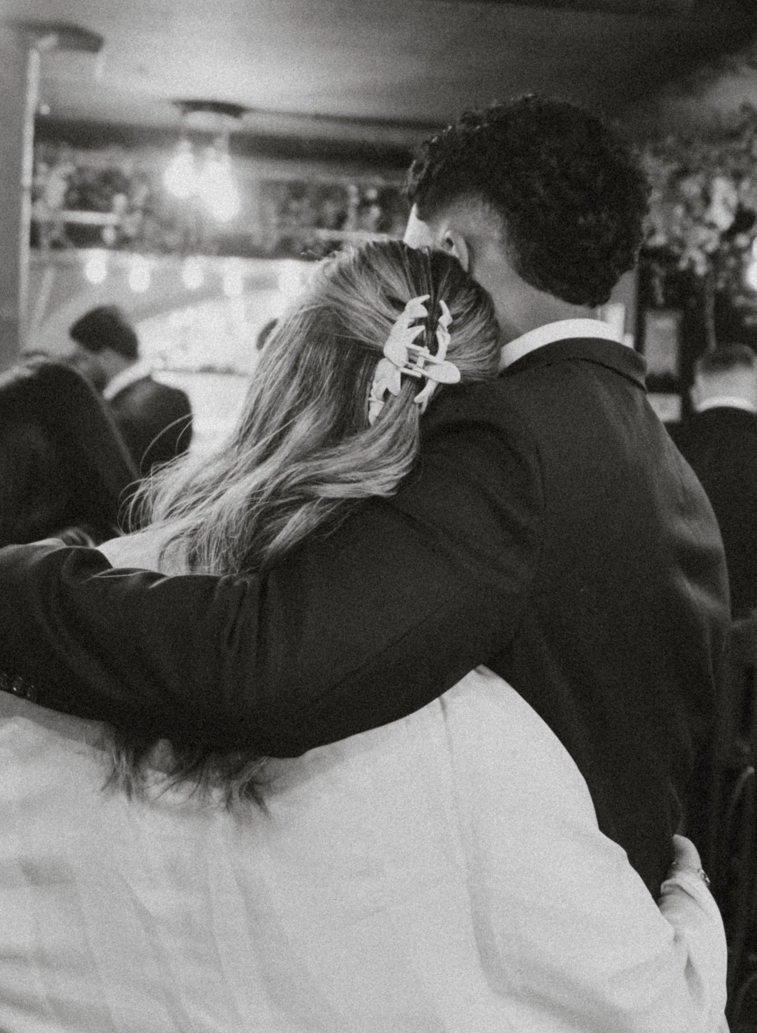 Black and white photo of a couple kissing, with the woman sitting on a surface and the man leaning into her, in an indoor setting with people in the background