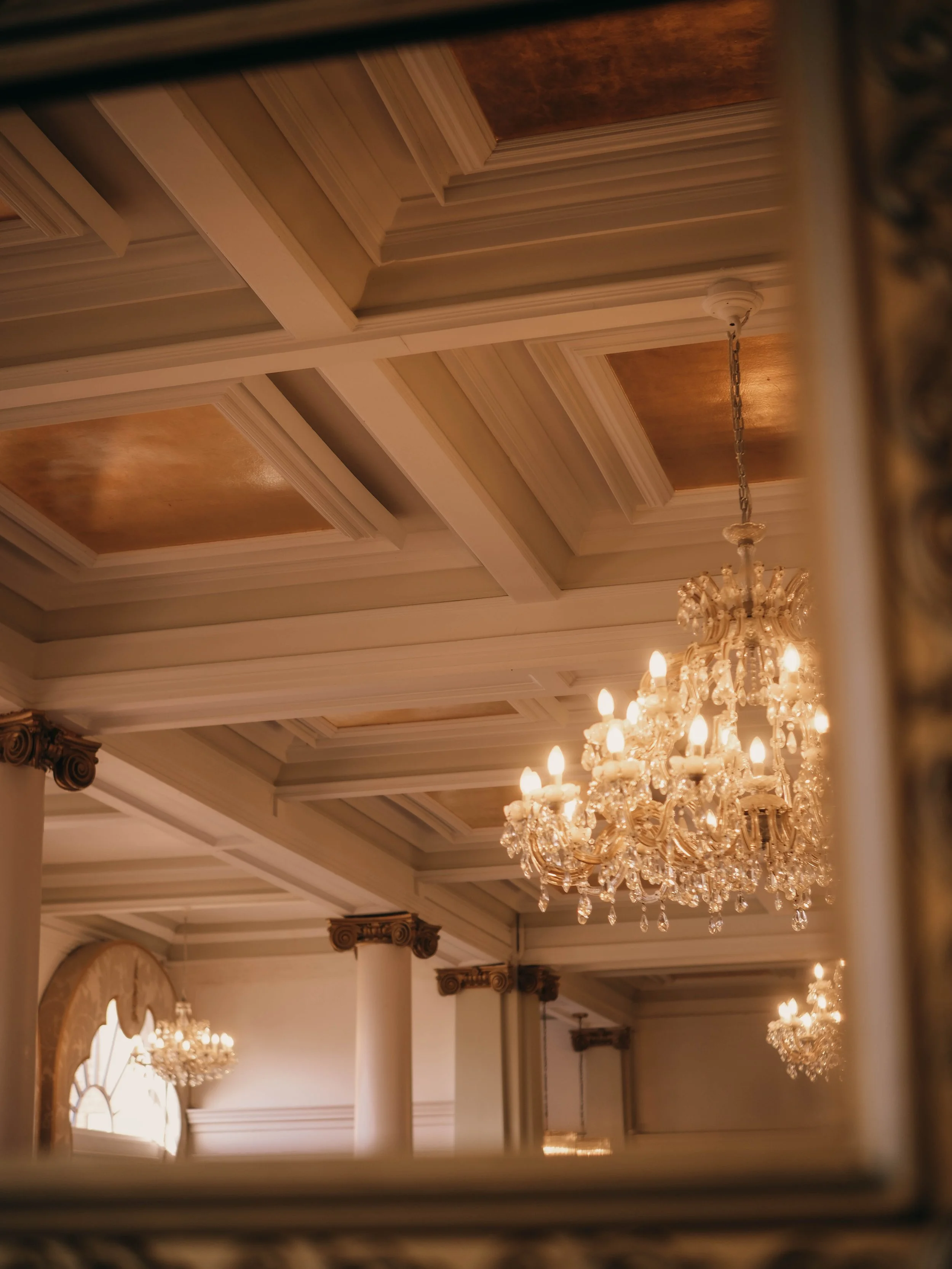A grand chandelier hanging from a decorated ceiling with ornate molding and warm lighting in an elegant interior space.