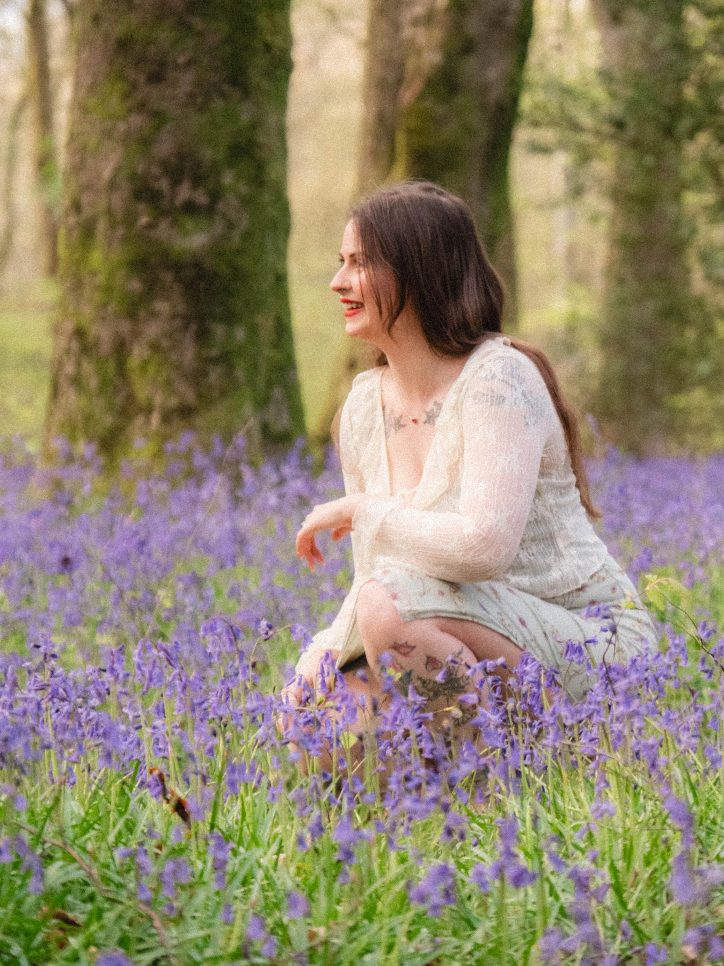 A woman with dark hair and tattoos, wearing a beige lace dress, is sitting in a field of purple flowers with trees in the background, smiling and looking to the side.
