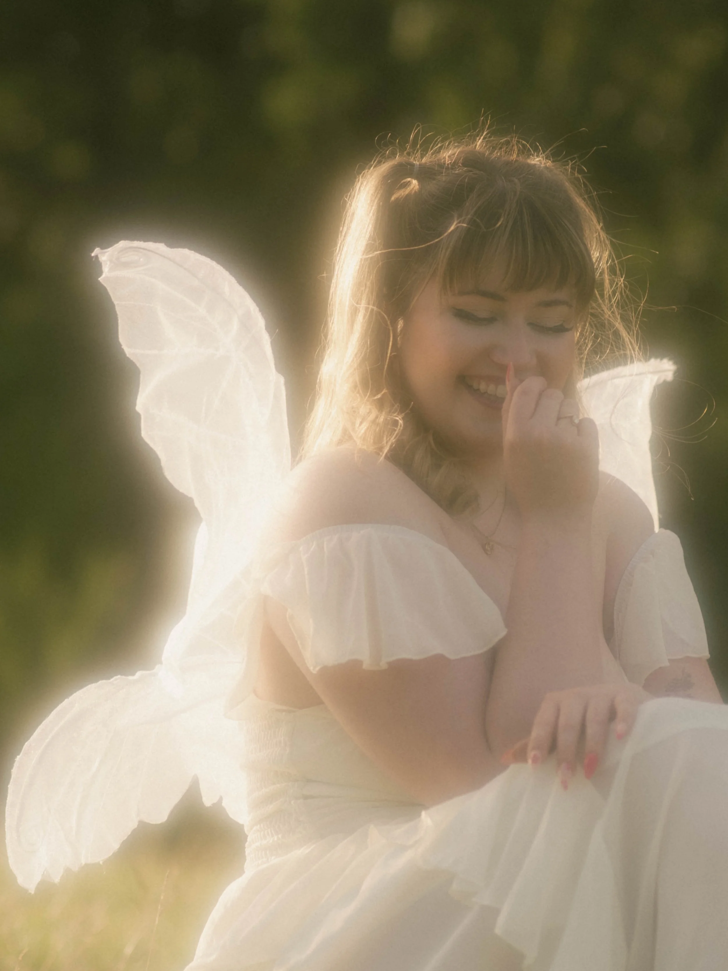 A woman dressed as a fairy or angel with white wings, wearing a white dress, smiling with her eyes closed and holding her nose, with light shining behind her and a blurred green background.