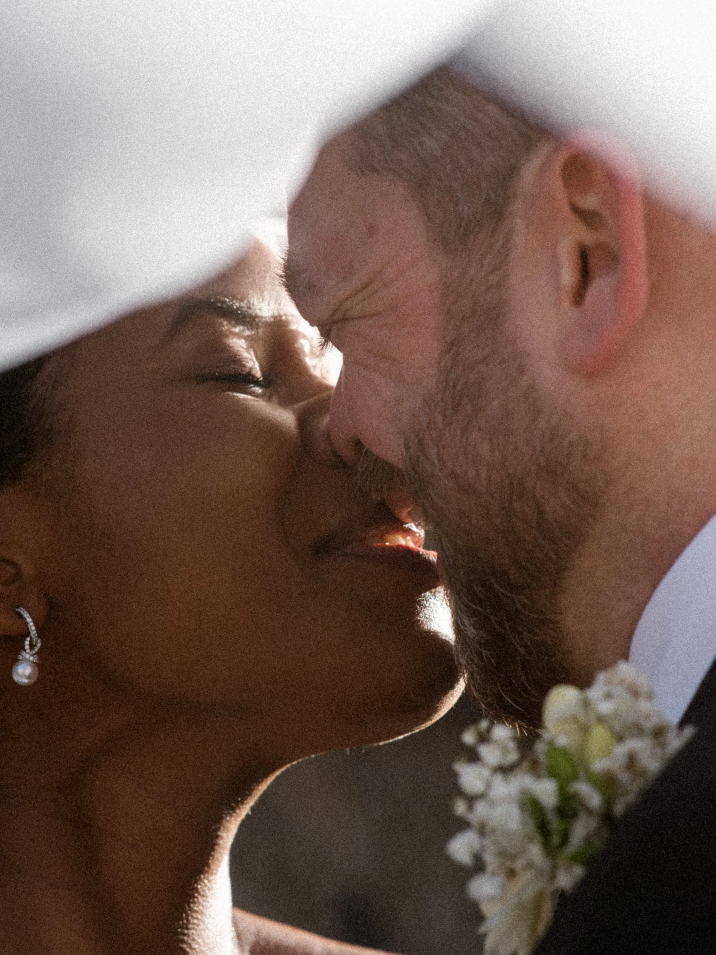 A close-up of a couple touching foreheads, with the woman wearing a pearl earring and the man having facial hair, near white flowers.