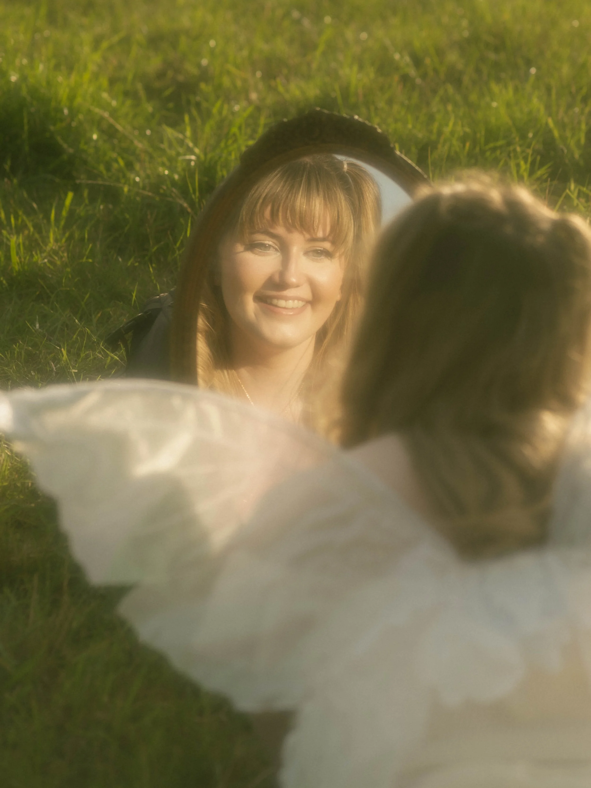 Reflection of a woman with red hair and bangs in a mirror outdoors, with grass in the background.