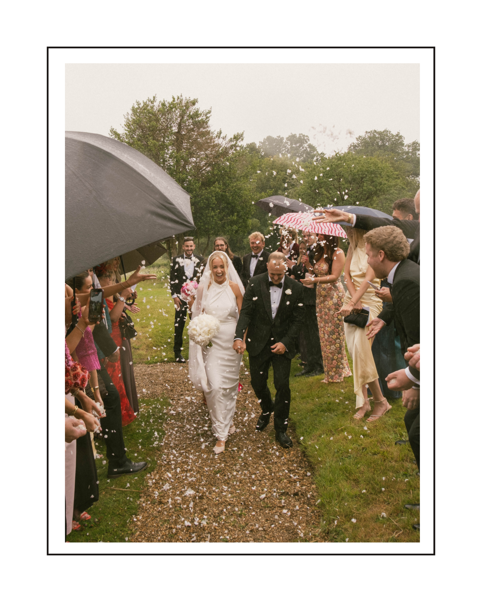 Bride and groom walking down an outdoor aisle during their wedding celebration, surrounded by guests holding umbrellas and throwing confetti.