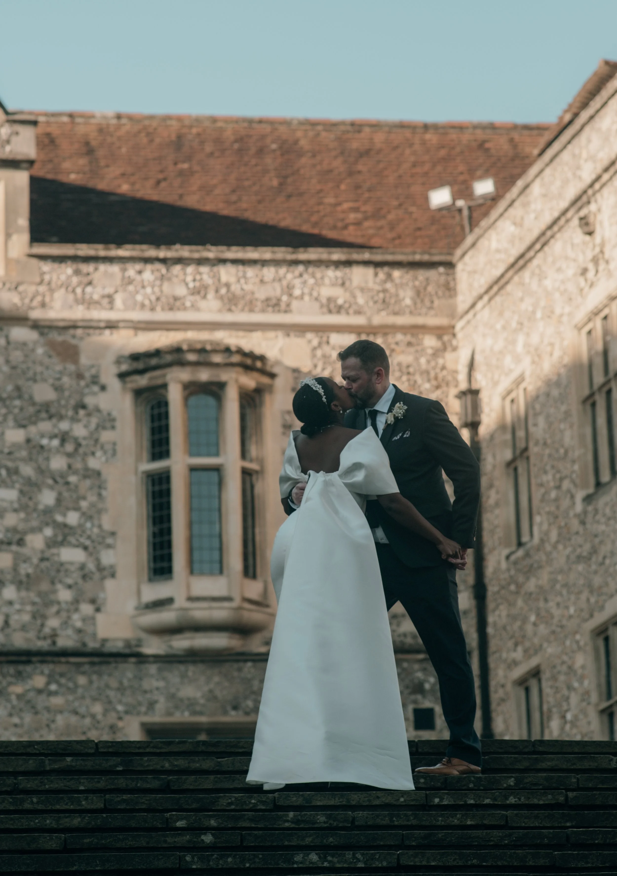 A bride and groom kiss on the steps of a historic stone building during their wedding, with the bride in a white gown and headpiece, and the groom in a dark suit.