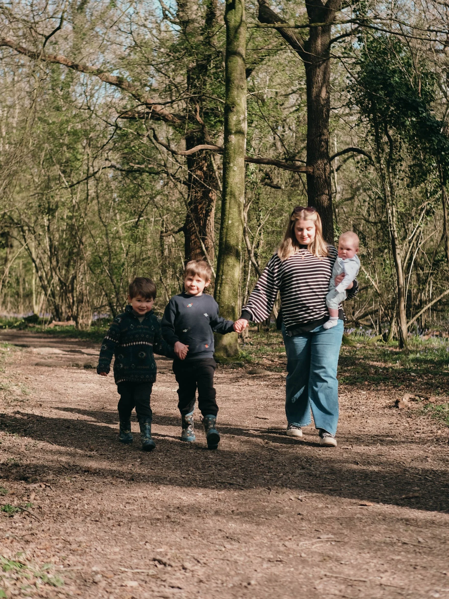 A woman and three children walking on a dirt trail in a wooded park during daytime.