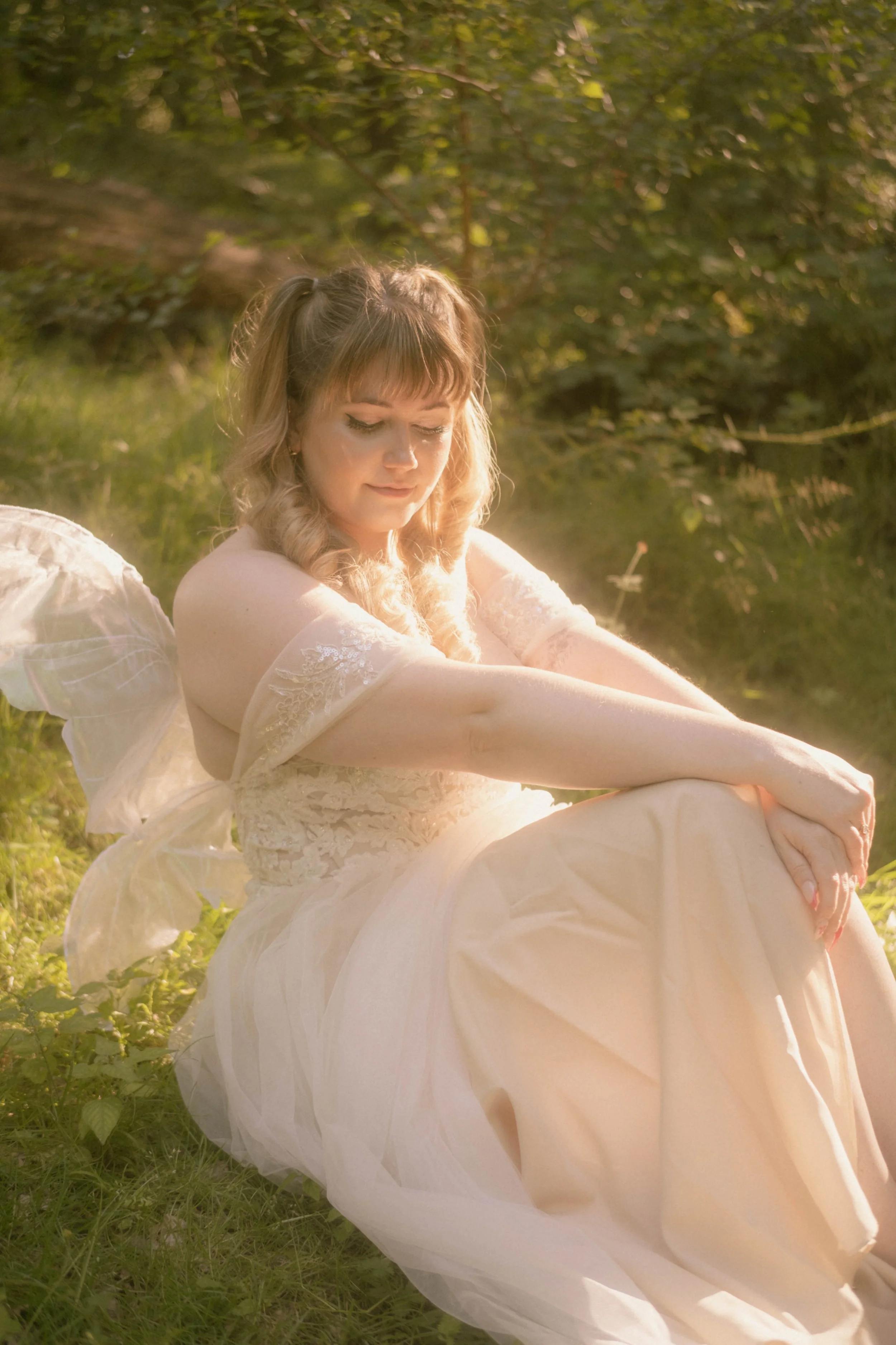 A young woman with blonde hair styled in curls, wearing a white dress and angel wings, sitting outdoors in a grassy area with trees, during golden hour sunlight.